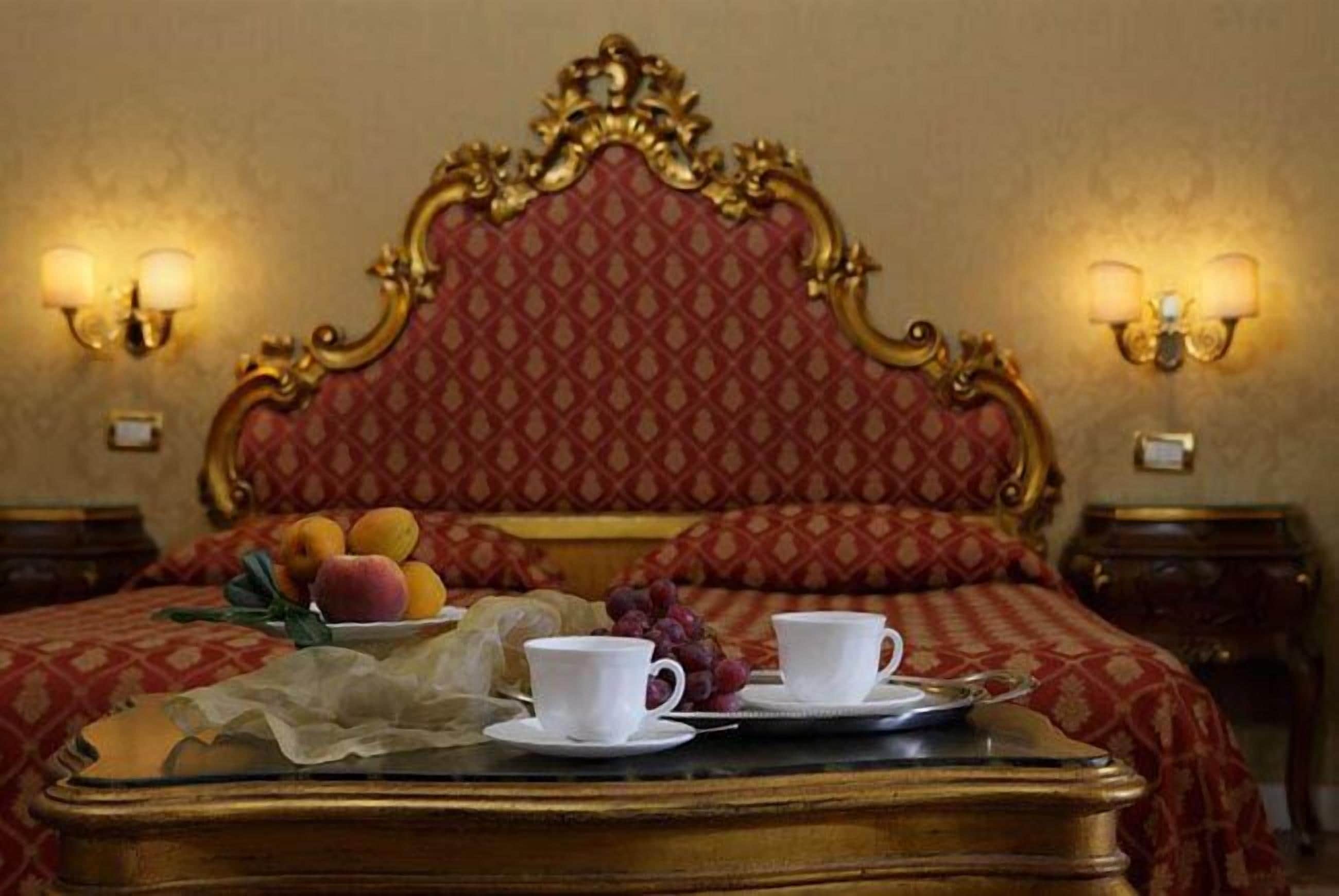 Un lit avec une tête de lit rouge et dorée, deux tables de chevet avec des lampes, et un plateau avec des fruits et des tasses posé sur le lit.