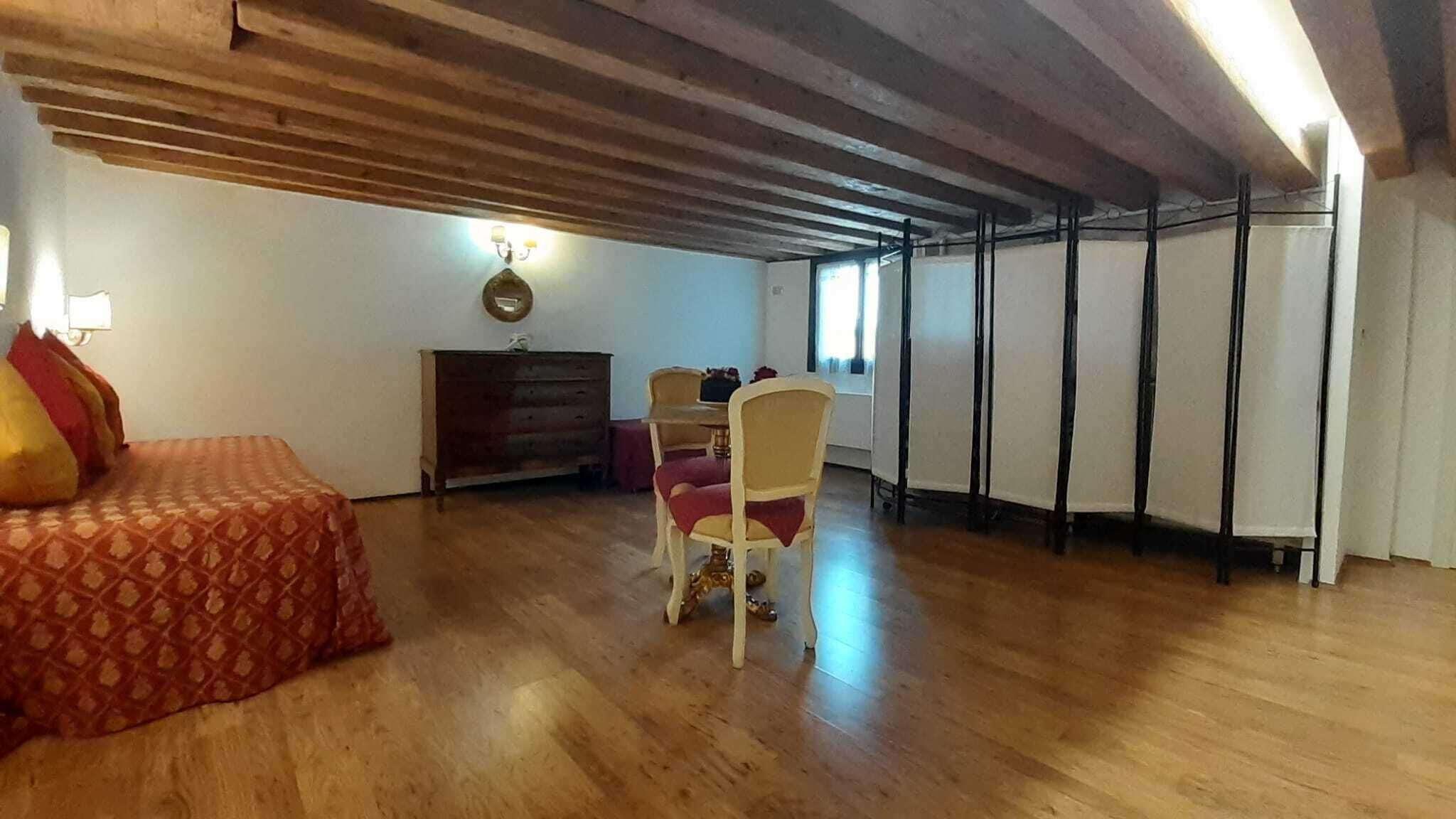 A room with a wooden ceiling, a bed with a patterned bedspread, a wooden dresser, a chair, and a table with a lamp.
