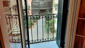 A view from a balcony with a metal railing, looking out onto a courtyard with potted plants and a building with arched windows.