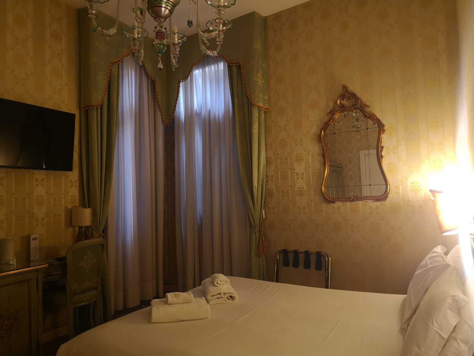 A hotel room with a bed, curtains, a chandelier, a mirror, a desk, and a chair.