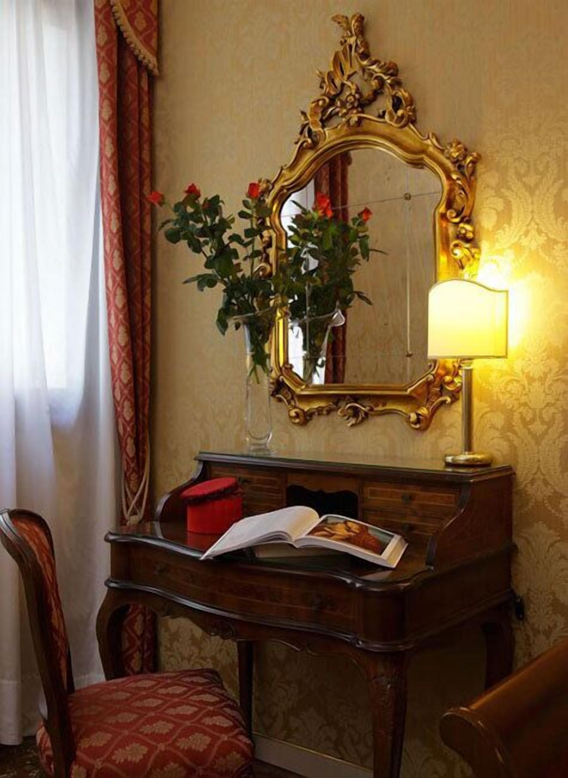 A room with a wooden desk, a mirror with an ornate frame, a vase with flowers, a lamp, and a chair.