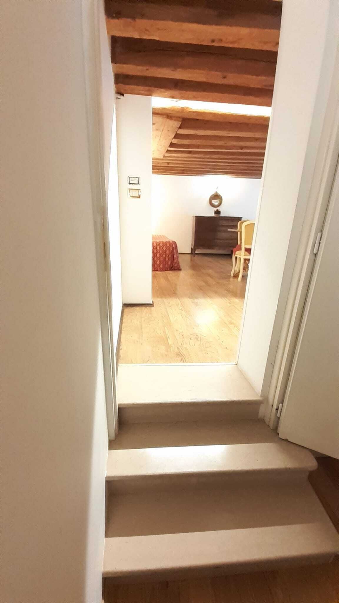 A staircase leading to a room with wooden flooring, a bed with a patterned bedspread, and a small table with a lamp.