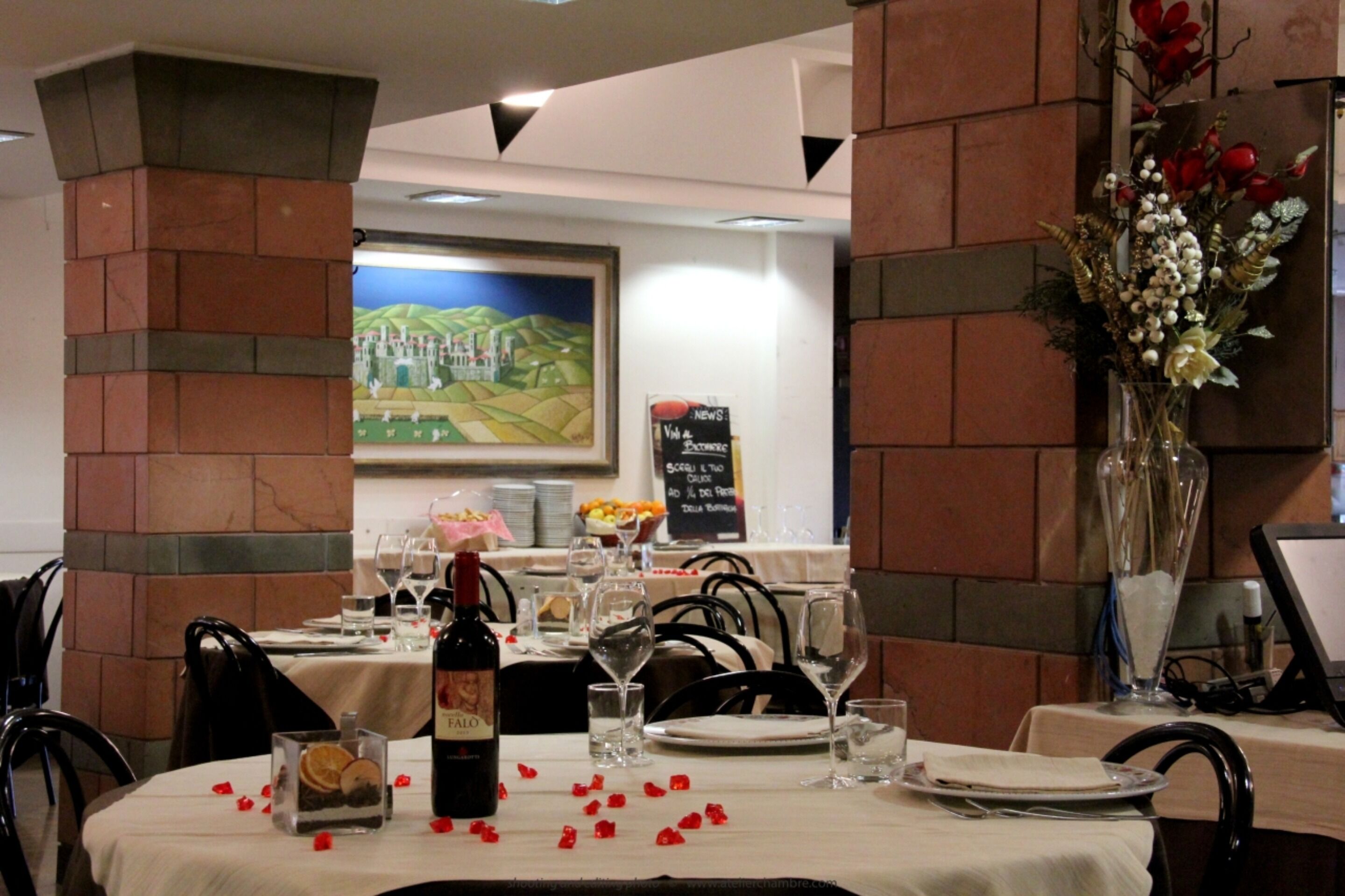 Un restaurant avec des piliers en briques, une table dressée avec des verres à vin, des assiettes et une bouteille de vin. Il y a un tableau au mur et un vase rempli de fleurs sur la table.