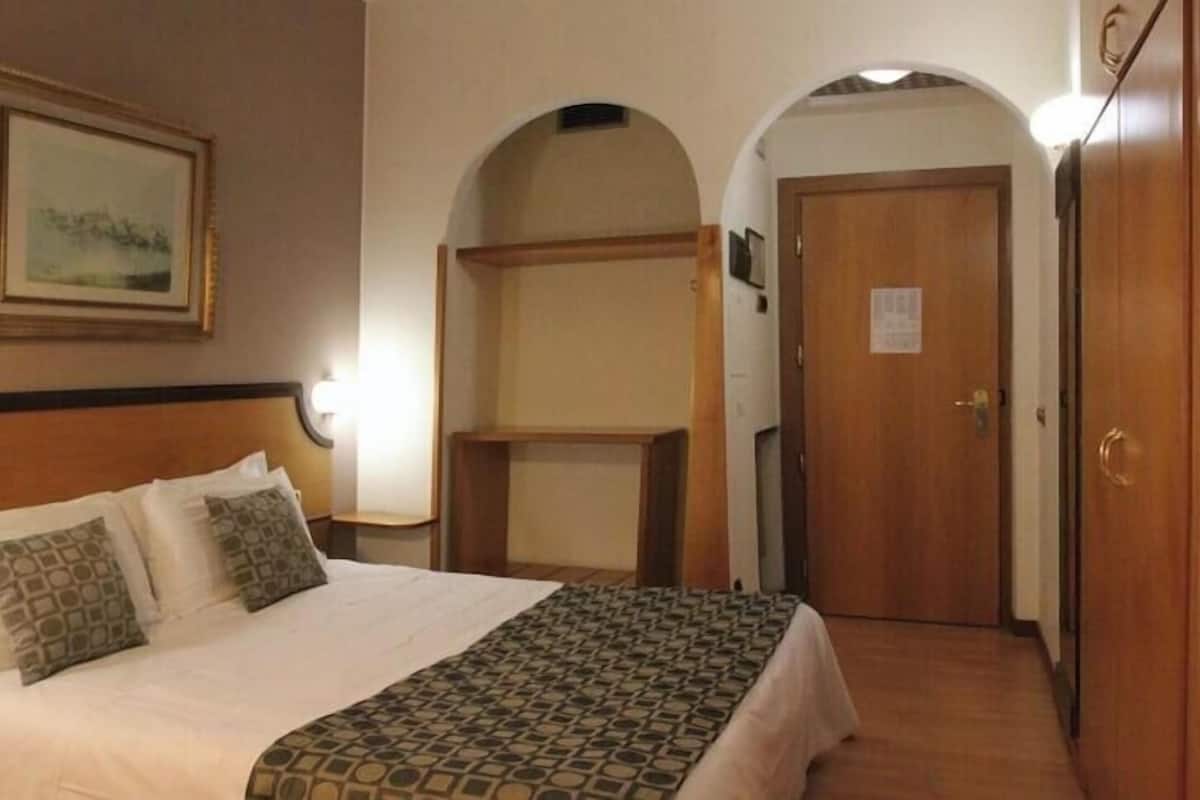 A hotel room with a bed, a bedside table, a lamp, a framed picture on the wall, and a door.