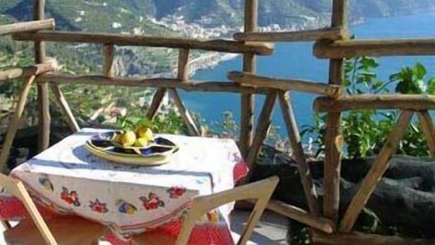 A table with a tablecloth and fruits, set against a backdrop of mountains and a body of water. - Giardino dei Limoni (Ravello)