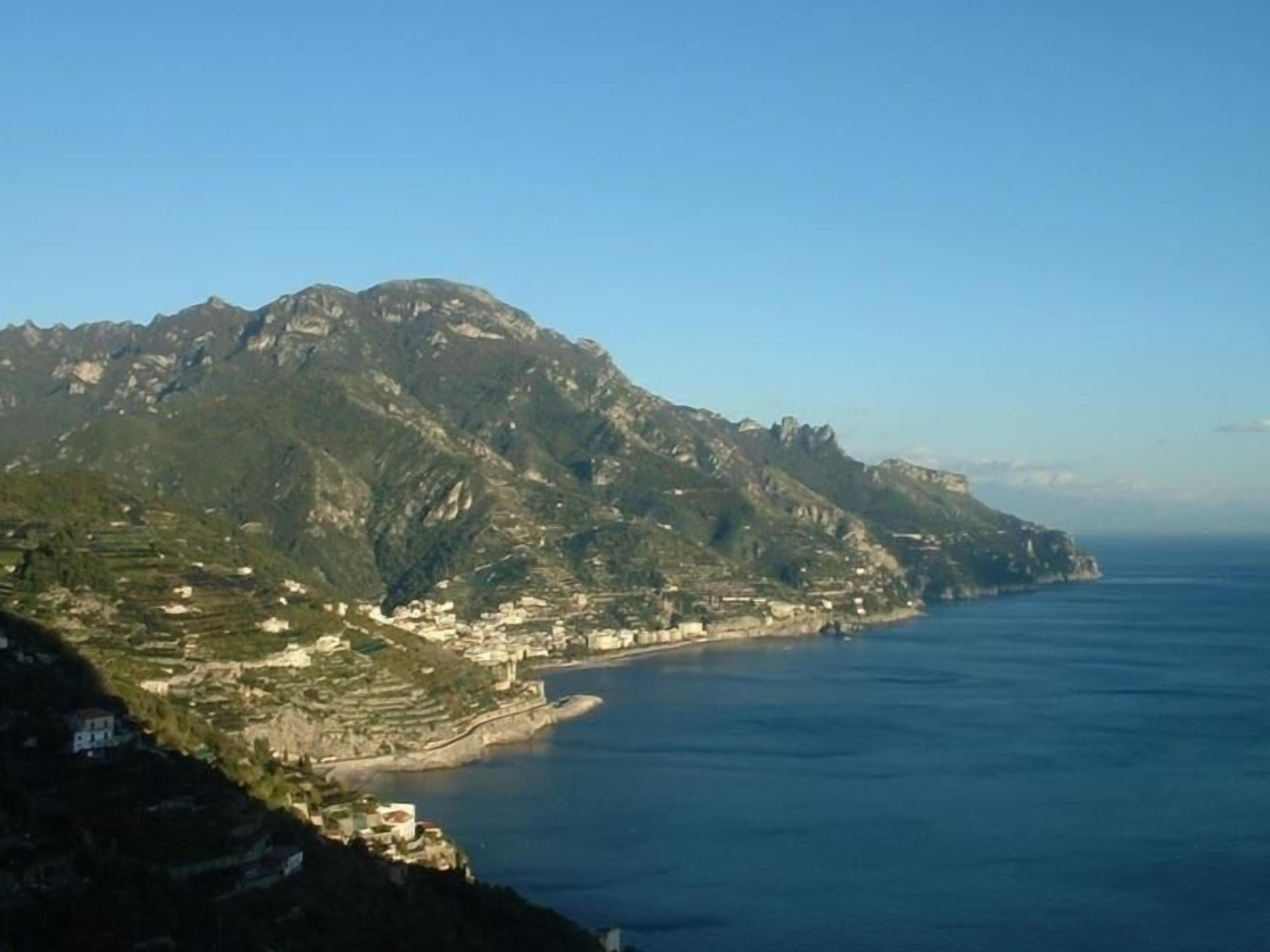 A coastal town with terraced buildings along the hillside, a clear view of the sea, and a prominent mountain in the background.