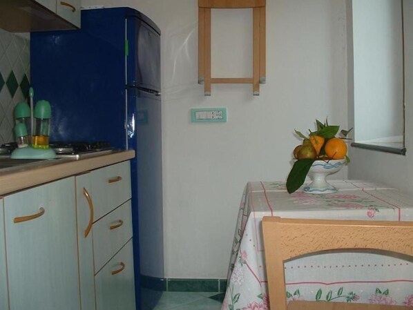 Une cuisine avec un réfrigérateur bleu, une table recouverte d’une nappe à motifs floraux et un vase contenant des oranges et des feuilles.