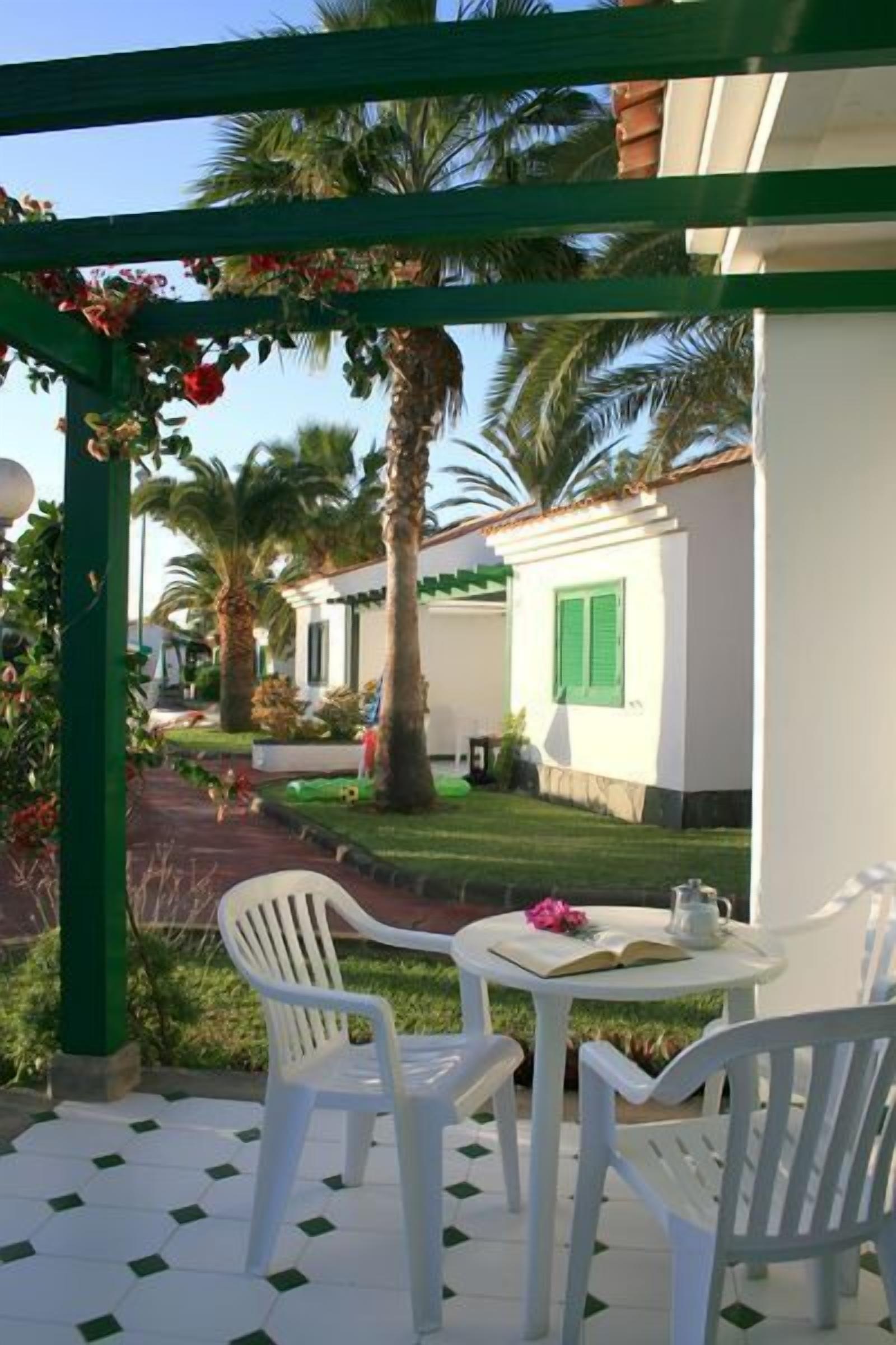 Um pátio com cadeiras e mesa de plástico branco, uma pérgula com flores vermelhas e um edifício branco com persianas verdes ao fundo.