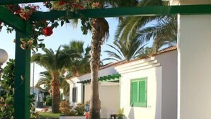 A patio with white plastic chairs and a table, a pergola with red flowers, and a white building with green shutters in the background. - Campo Golf Bungalows (San Bartolomé de Tirajana)