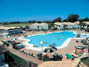 Una gran piscina rodeada de sillas de descanso y sombrillas, con vista al océano y un edificio con techo de tejas al fondo.