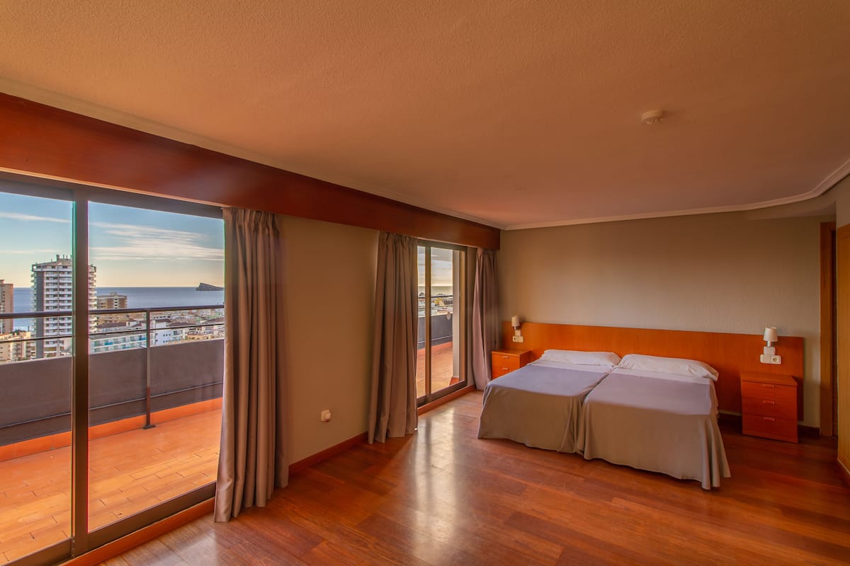 A hotel room with a balcony, a double bed, a nightstand, and a view of the city and sea.