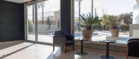 Amplia habitación con ventanas grandes, un área de descanso con sillas y una mesa, y vista a una piscina y palmeras fuera.