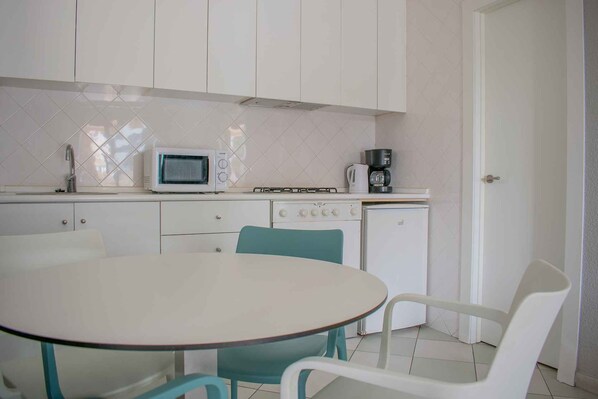 Cocina compacta con armarios blancos, microondas, estufa y cafetera. Hay una pequeña mesa de comedor con sillas.