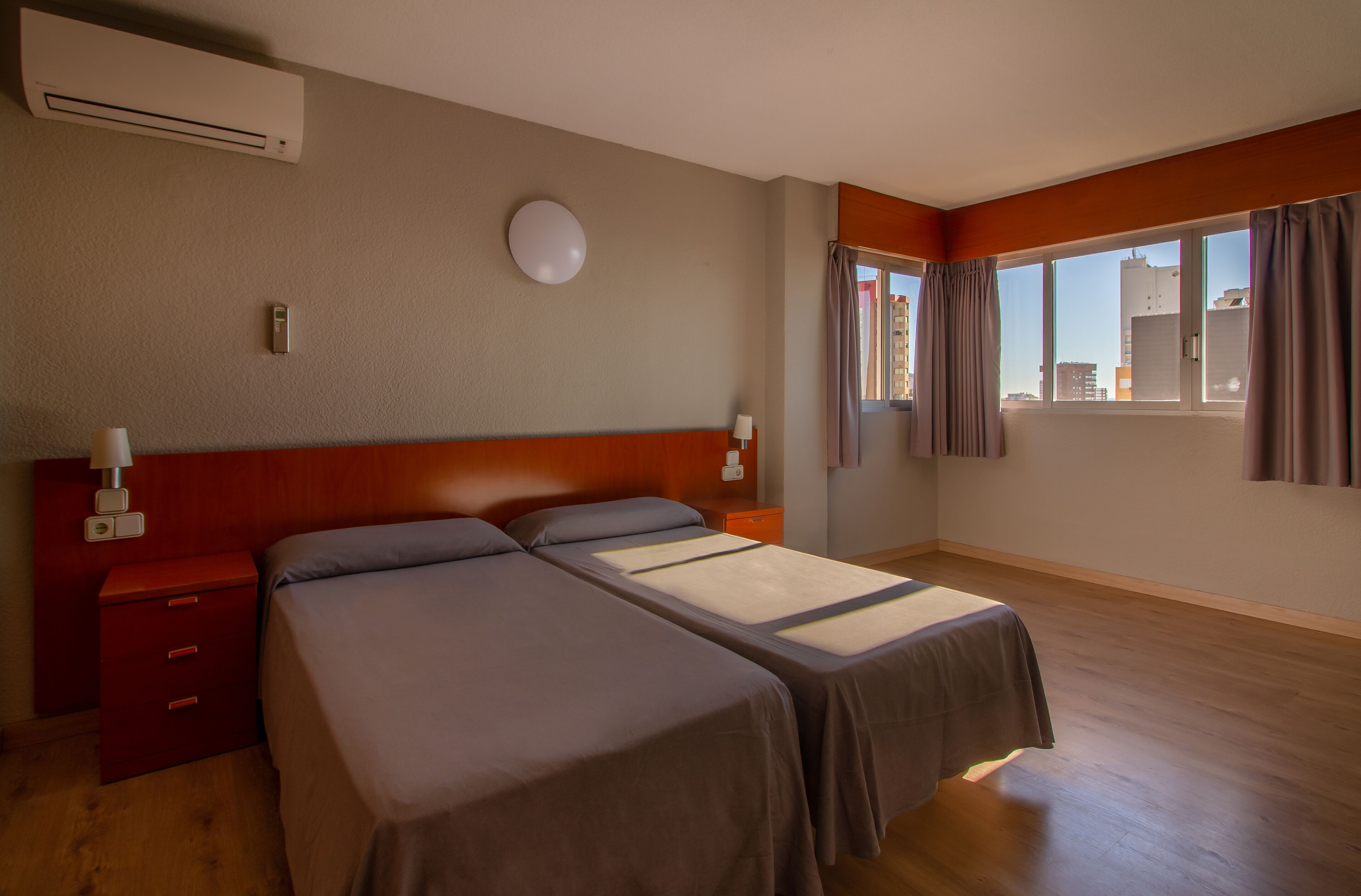 Een hotelkamer met twee bedden, een houten vloer, een raam met gordijnen en een wandairconditioner.