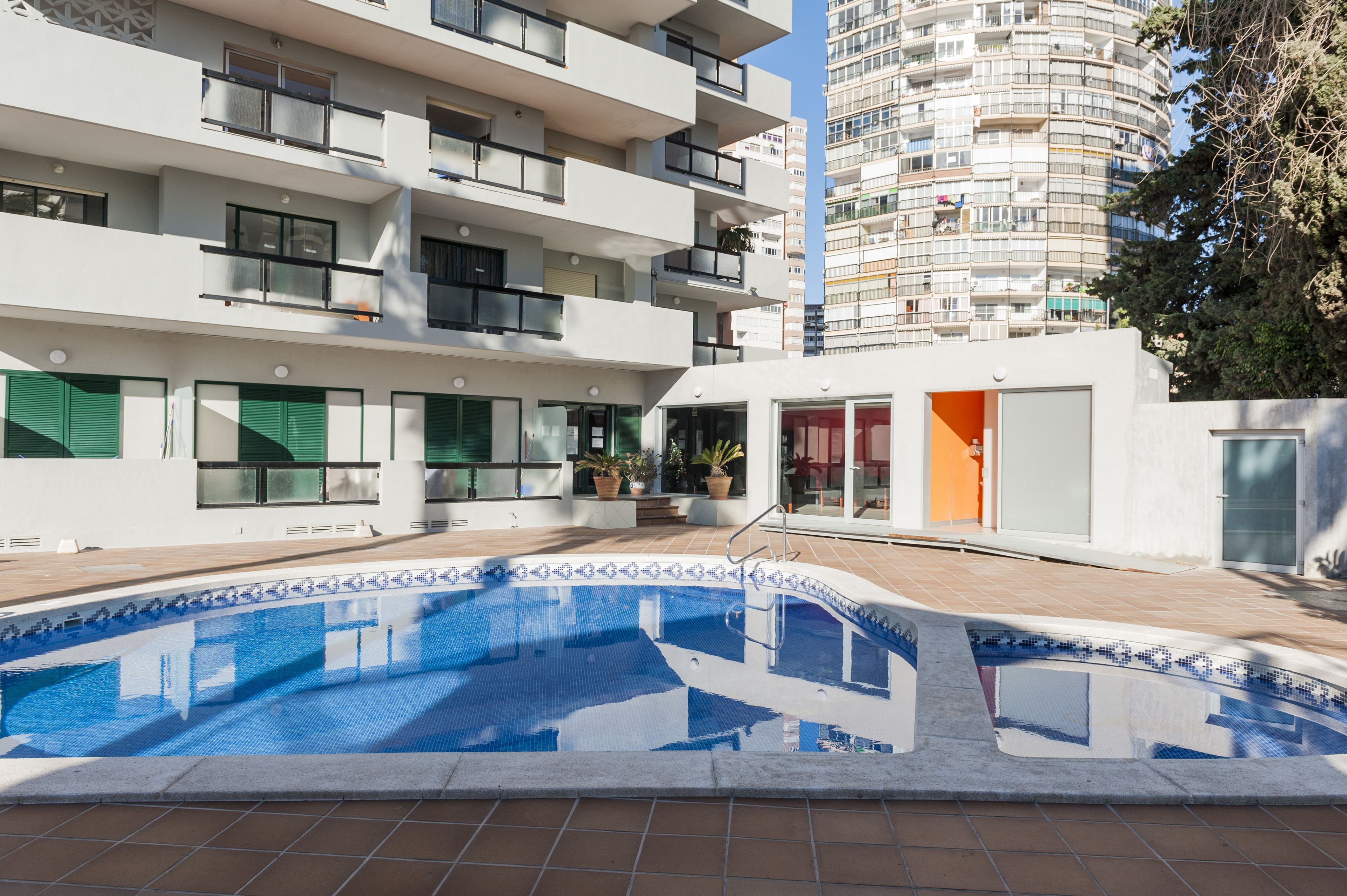 A modern building with a swimming pool, balconies, and a view of a tall residential building in the background.