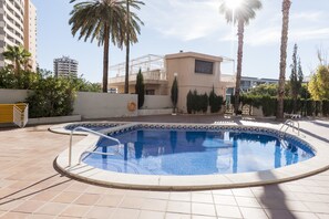 Una piscina con borde de baldosas, rodeada de palmeras y un edificio con balcón.