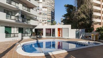Un área de piscina con terraza de baldosas, una piscina con diseño azul y blanco y un edificio con varios balcones y ventanas.
