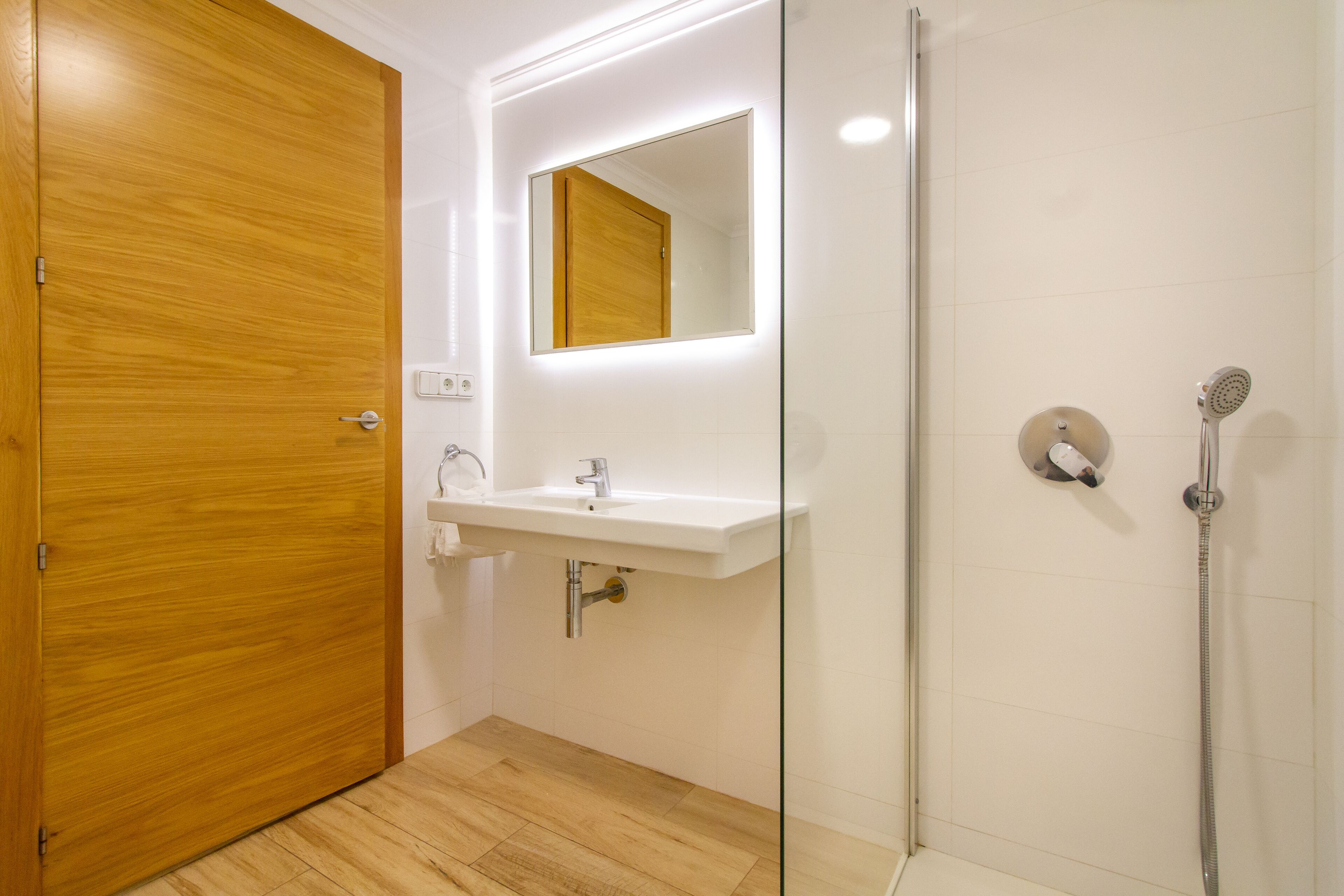 Un baño moderno con una puerta de madera, un lavabo blanco, un espejo y una ducha.