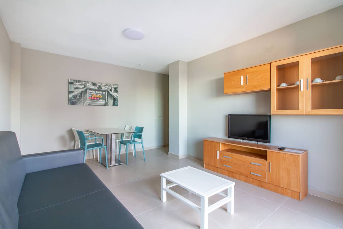 A modern living room with a sofa, a coffee table, a TV on a wooden cabinet, and a dining area with chairs and a table.