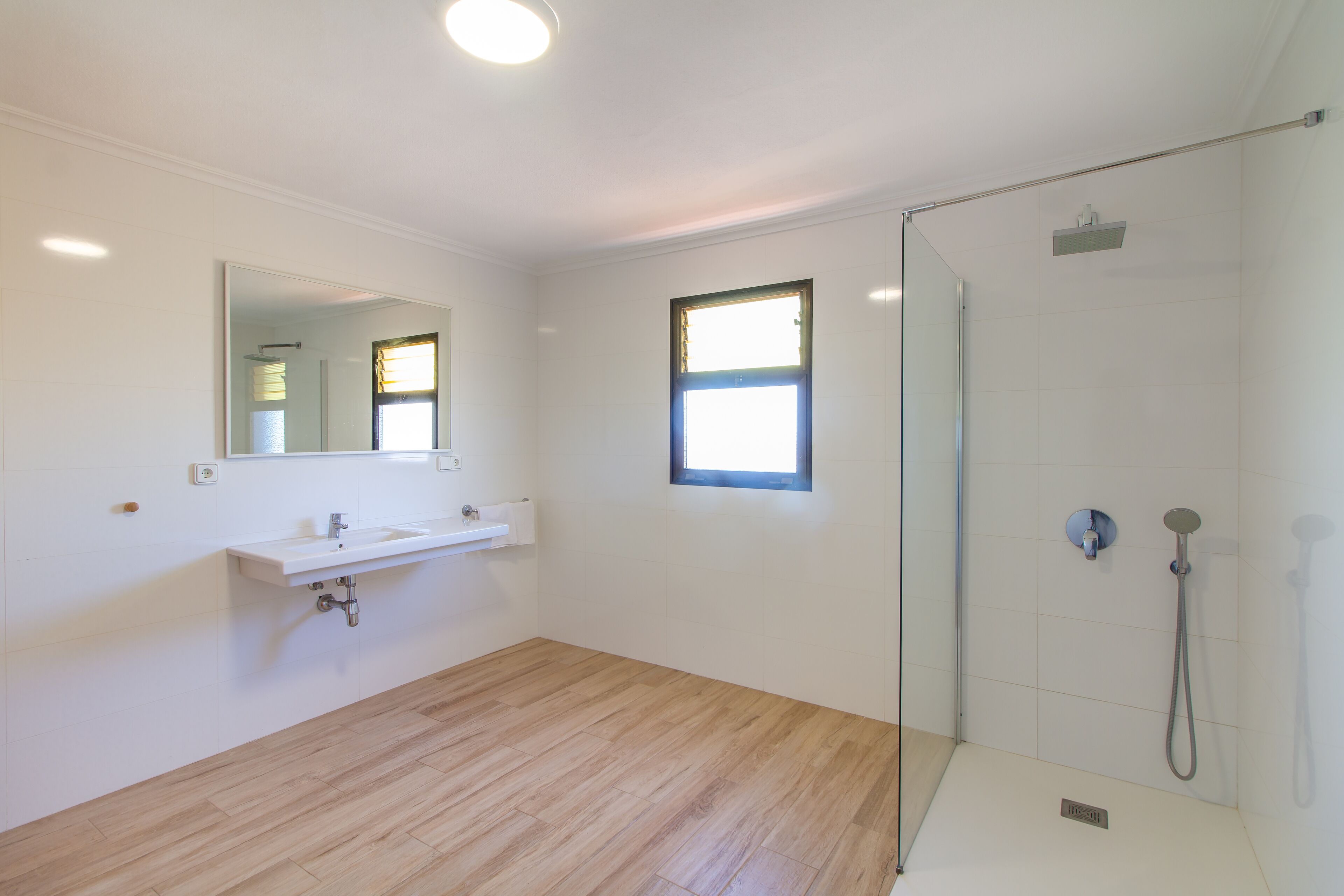 Une salle de bain avec un plancher en bois, un lavabo blanc, un miroir, une douche avec une porte en verre et une petite fenêtre.