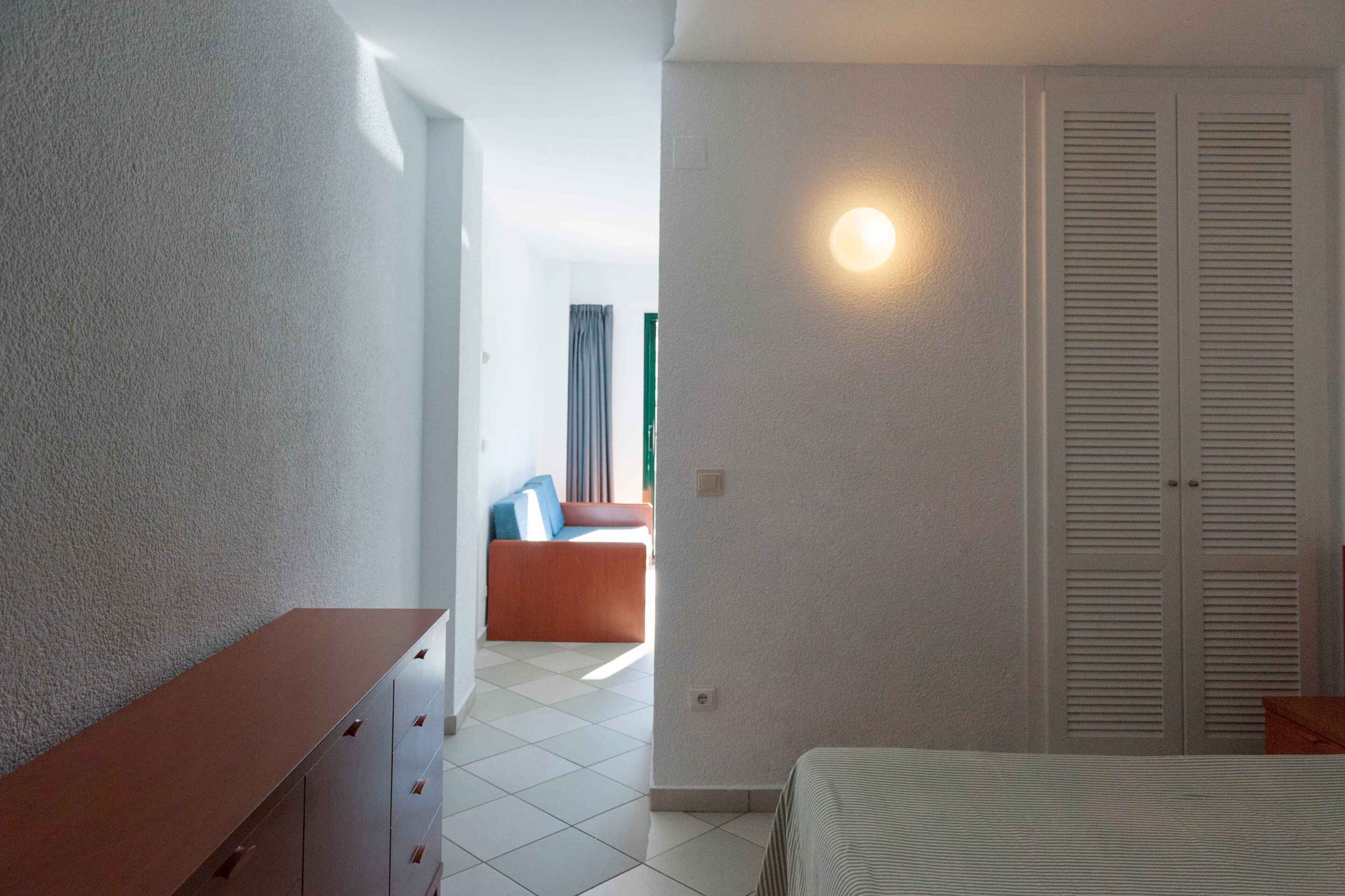 Une chambre à coucher comprenant un lit, une commode, un placard et donnant sur une autre pièce où se trouvent un canapé et des rideaux.