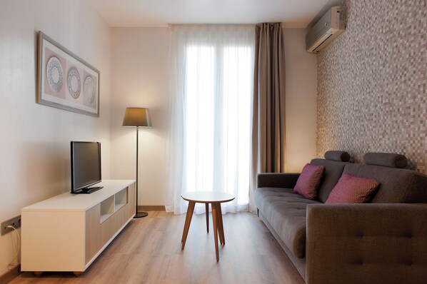 Ein modernes Wohnzimmer mit einem Flachbildfernseher, einem kleinen runden Tisch, einer Couch mit gemusterten Kissen und einem großen Fenster mit durchsichtigen Vorhängen.