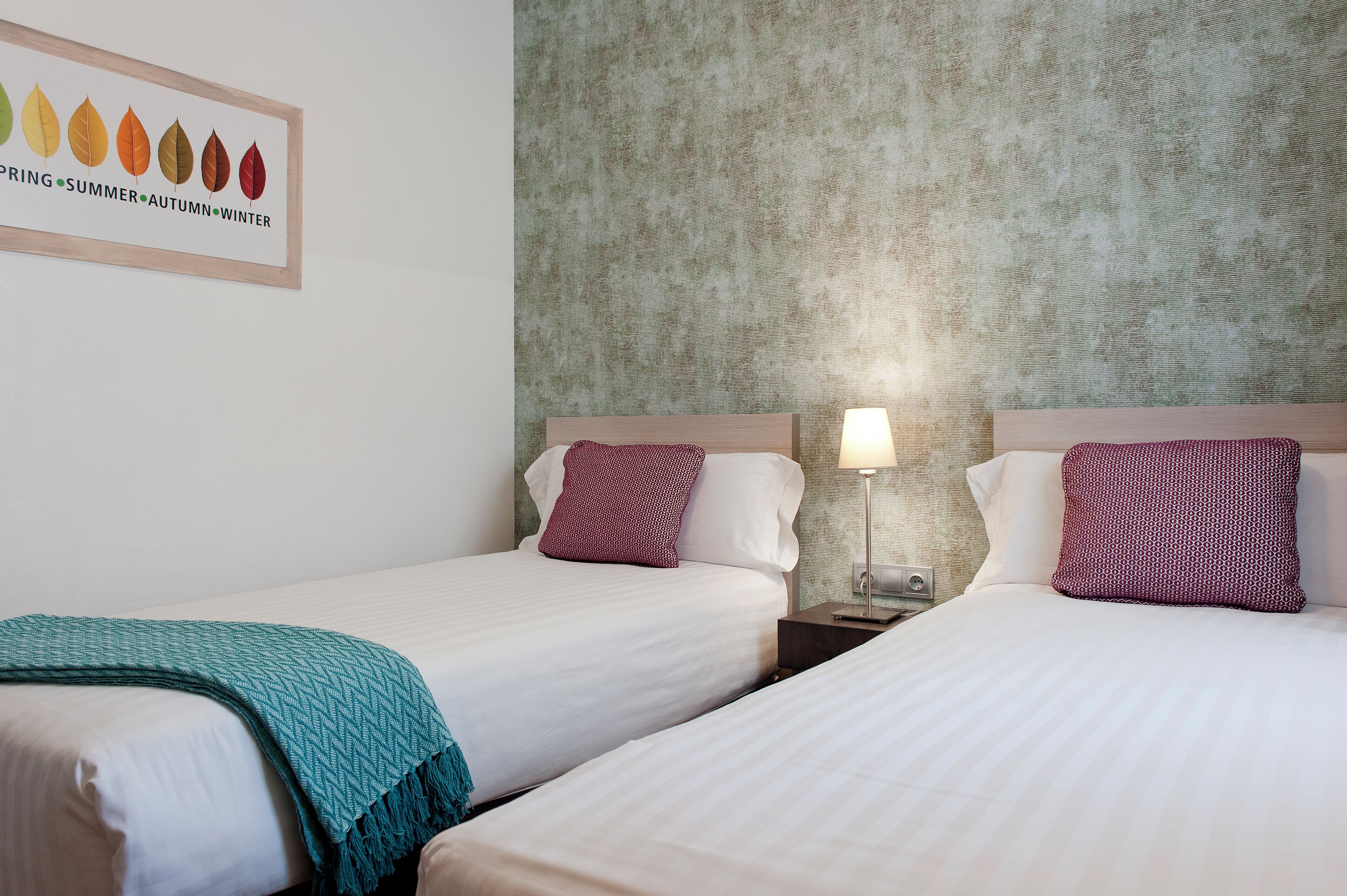 Habitación de hotel con dos camas, una pared con diseño de hojas y una mesita de noche con una lámpara.