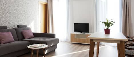 Una sala de estar moderna con un sofá gris, una mesita redonda pequeña, un televisor sobre un mueble de madera y una planta en maceta.