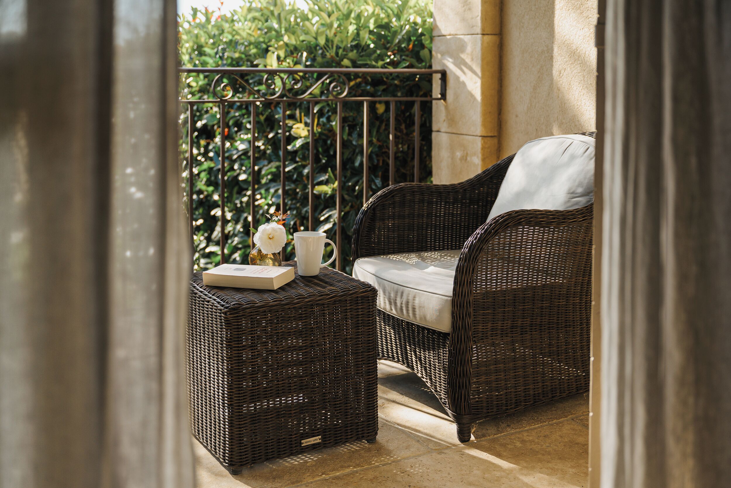 A wicker chair and ottoman on a balcony with a railing, a white cushion, and a small table with a book and a white flower.