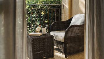 A wicker chair and ottoman on a balcony with a railing, a white cushion, and a small table with a book and a white flower.