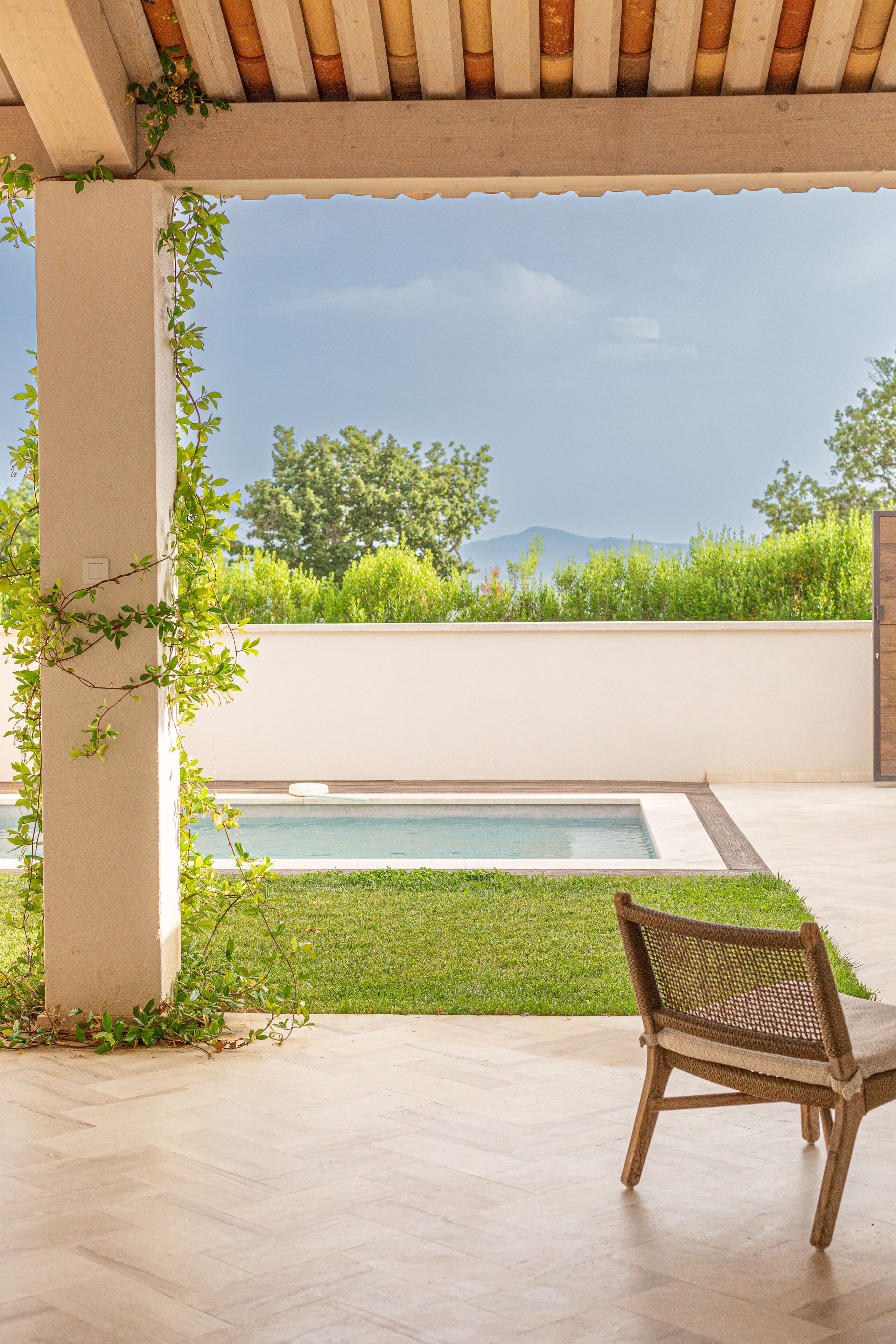 A covered patio with a pool, a chair, and a view of greenery and mountains.