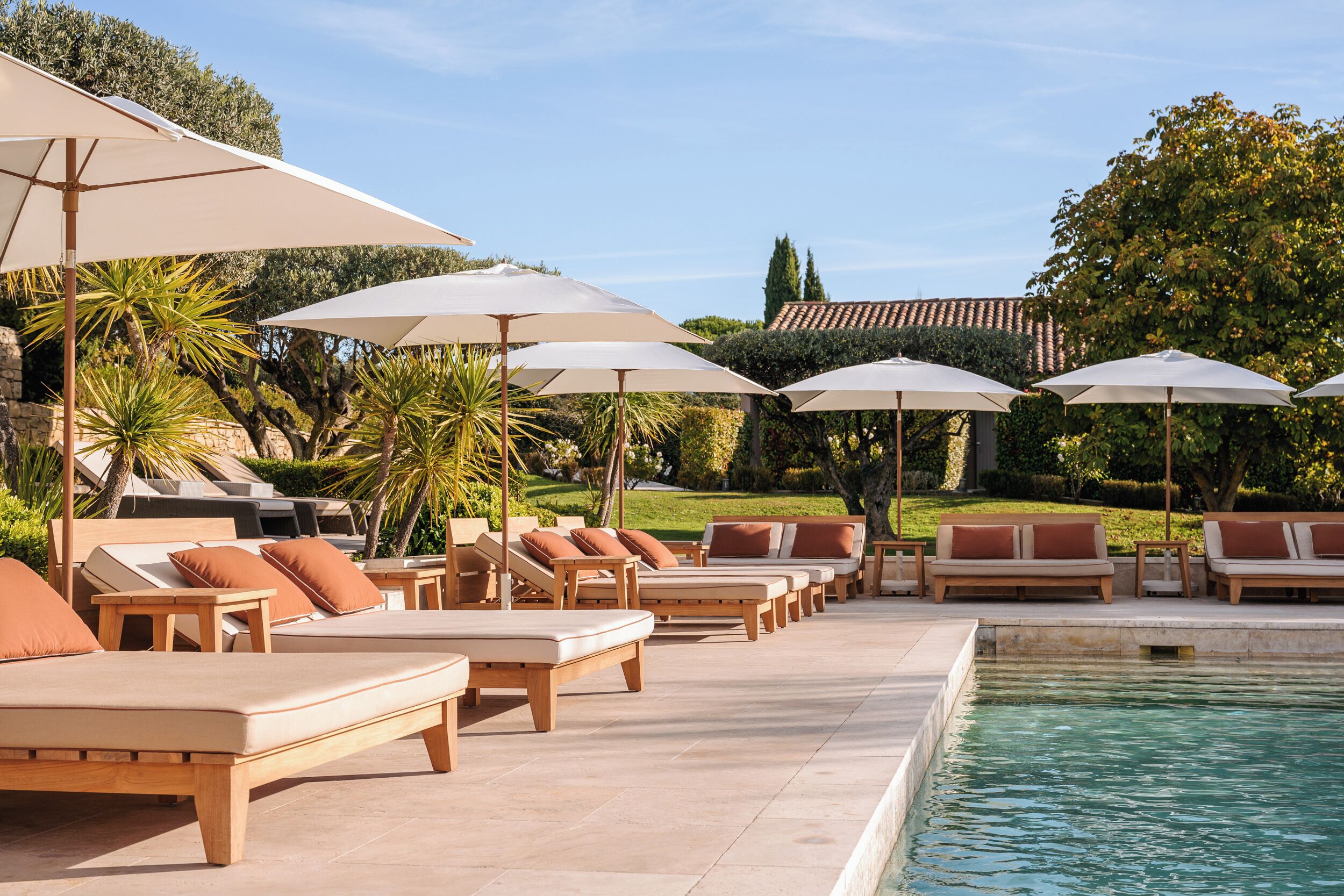 Un área junto a la piscina con sillones de descanso y sombrillas, rodeada de vegetación y un cielo despejado.