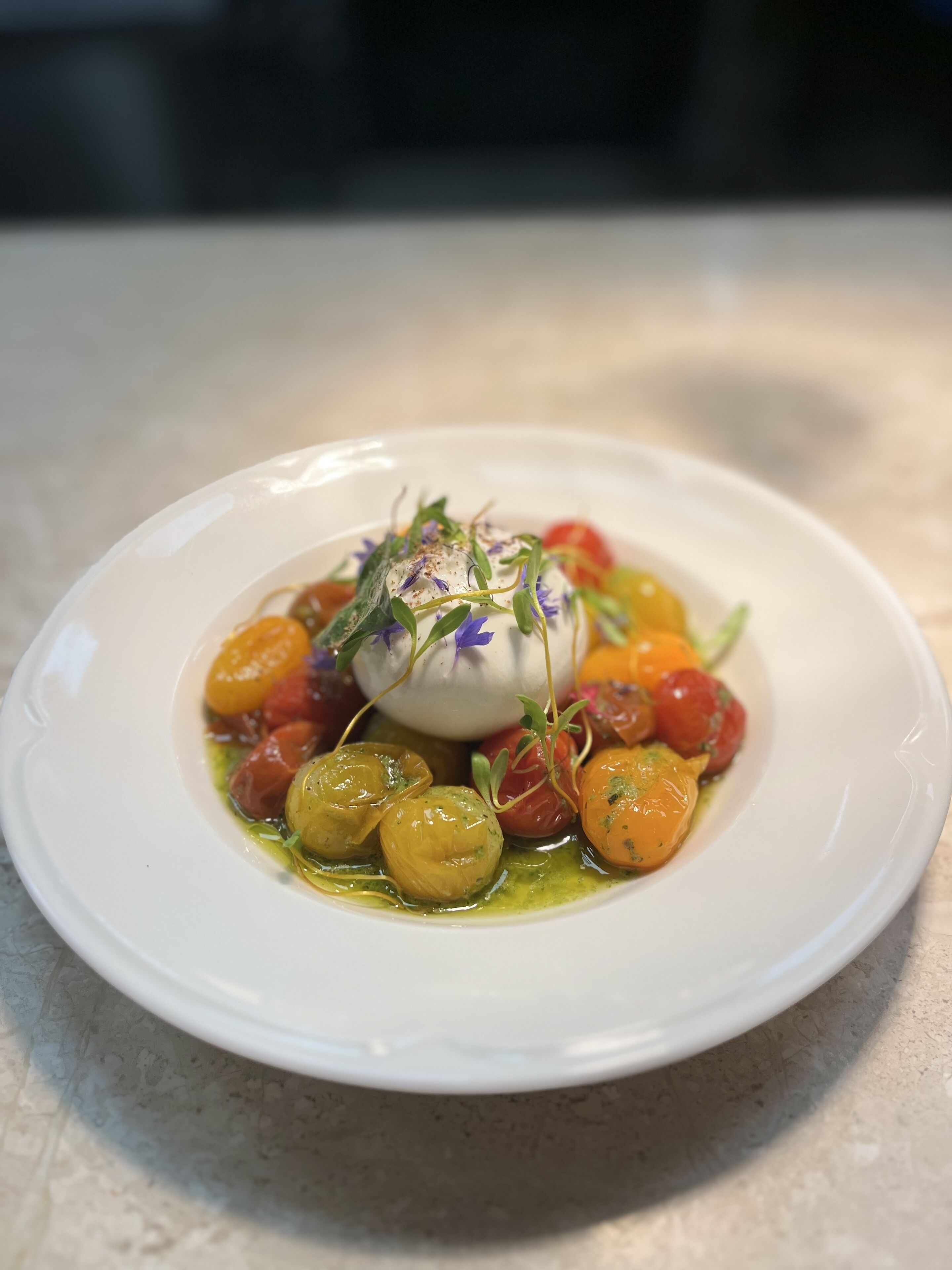 Une assiette blanche contenant une salade composée de billes de mozzarella, de tomates cerises et de jeunes pousses, le tout agrémenté d’une fleur violette.