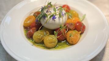 Une assiette blanche contenant une salade composée de billes de mozzarella, de tomates cerises et de jeunes pousses, le tout agrémenté d’une fleur violette.