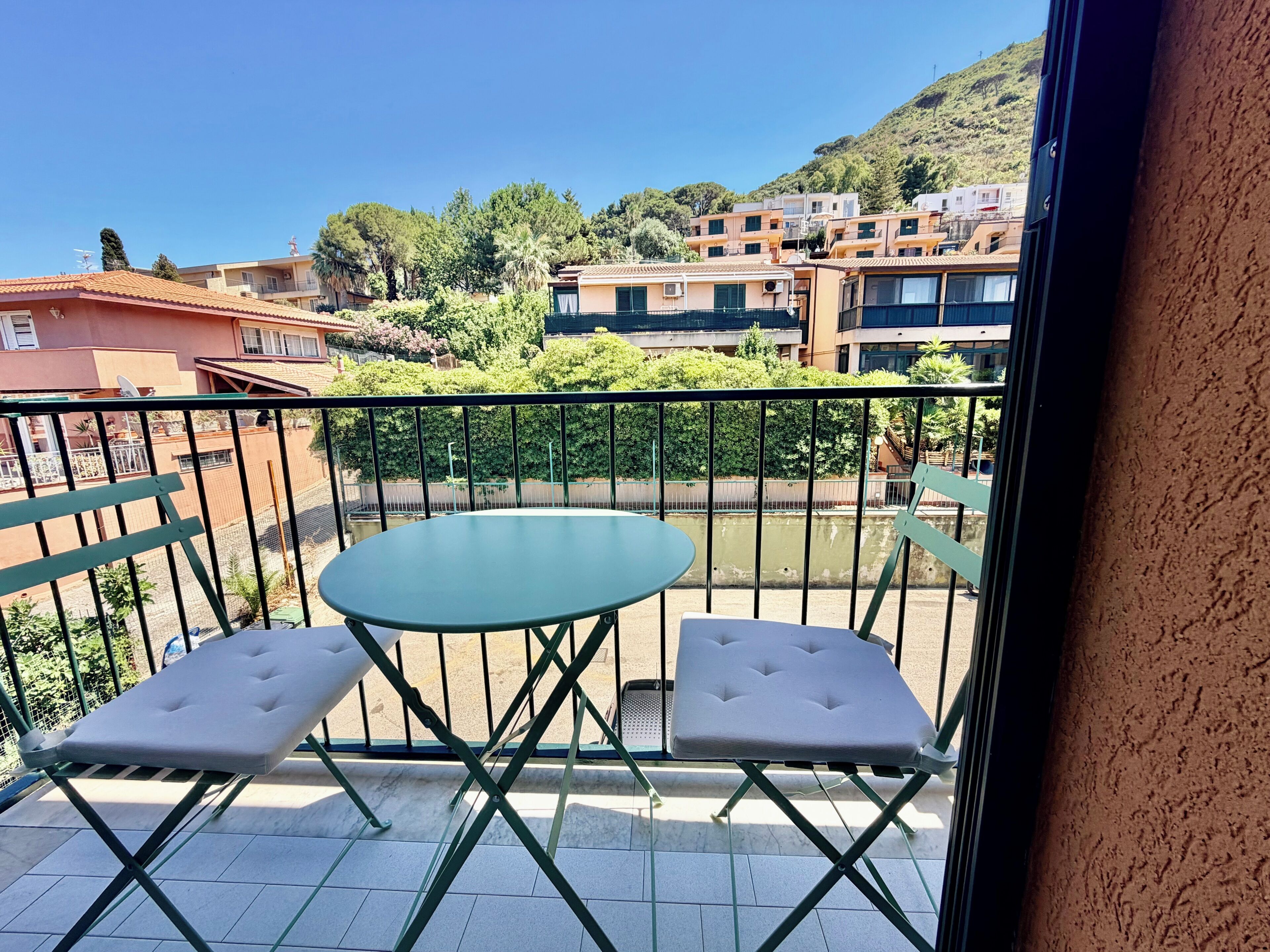 Un balcon avec une table et deux fauteuils confortables, donnant sur un quartier résidentiel avec des immeubles et de la verdure.
