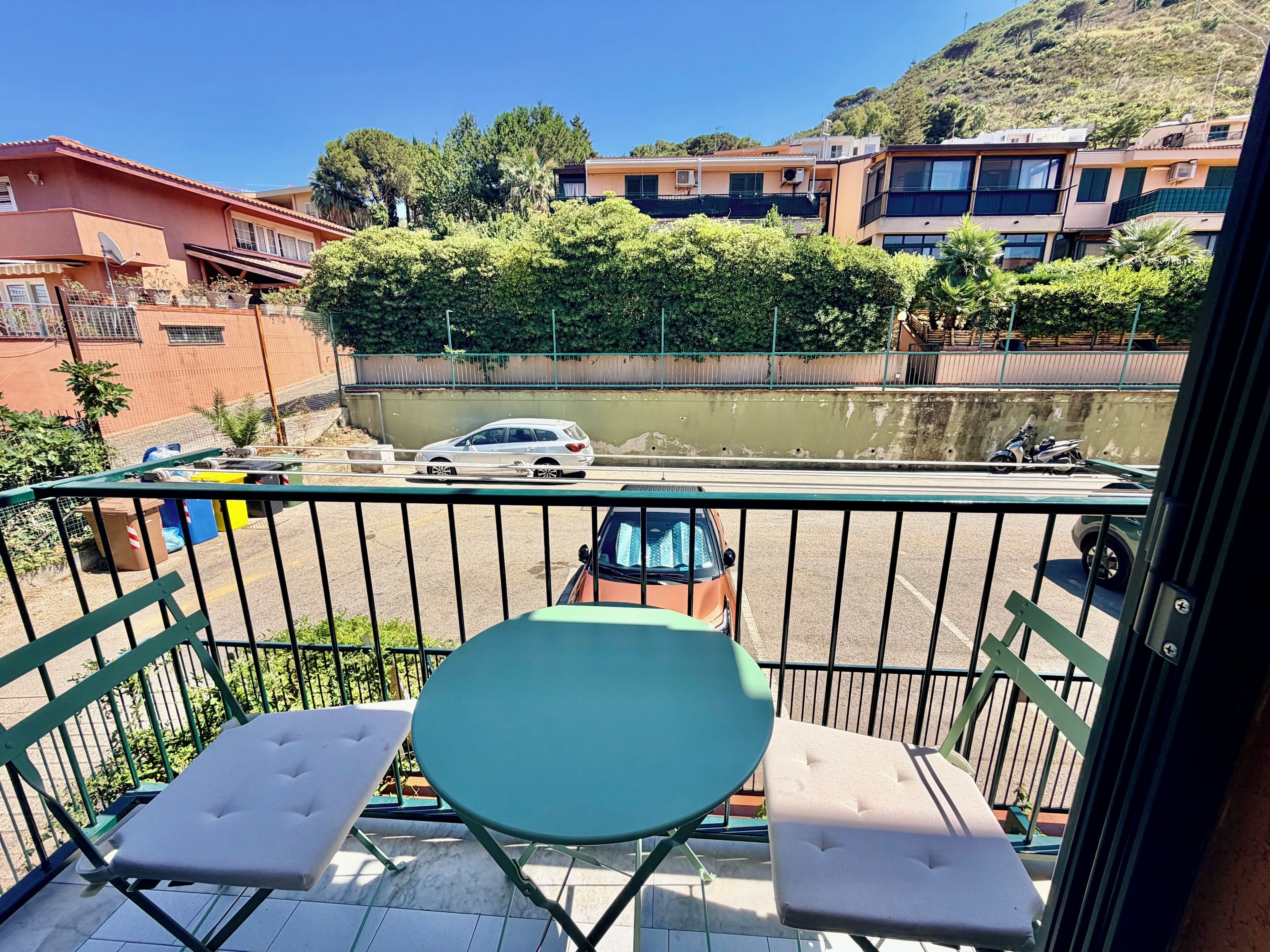 A balcony with two cushioned chairs and a table, overlooking a road with vehicles and buildings in the background.