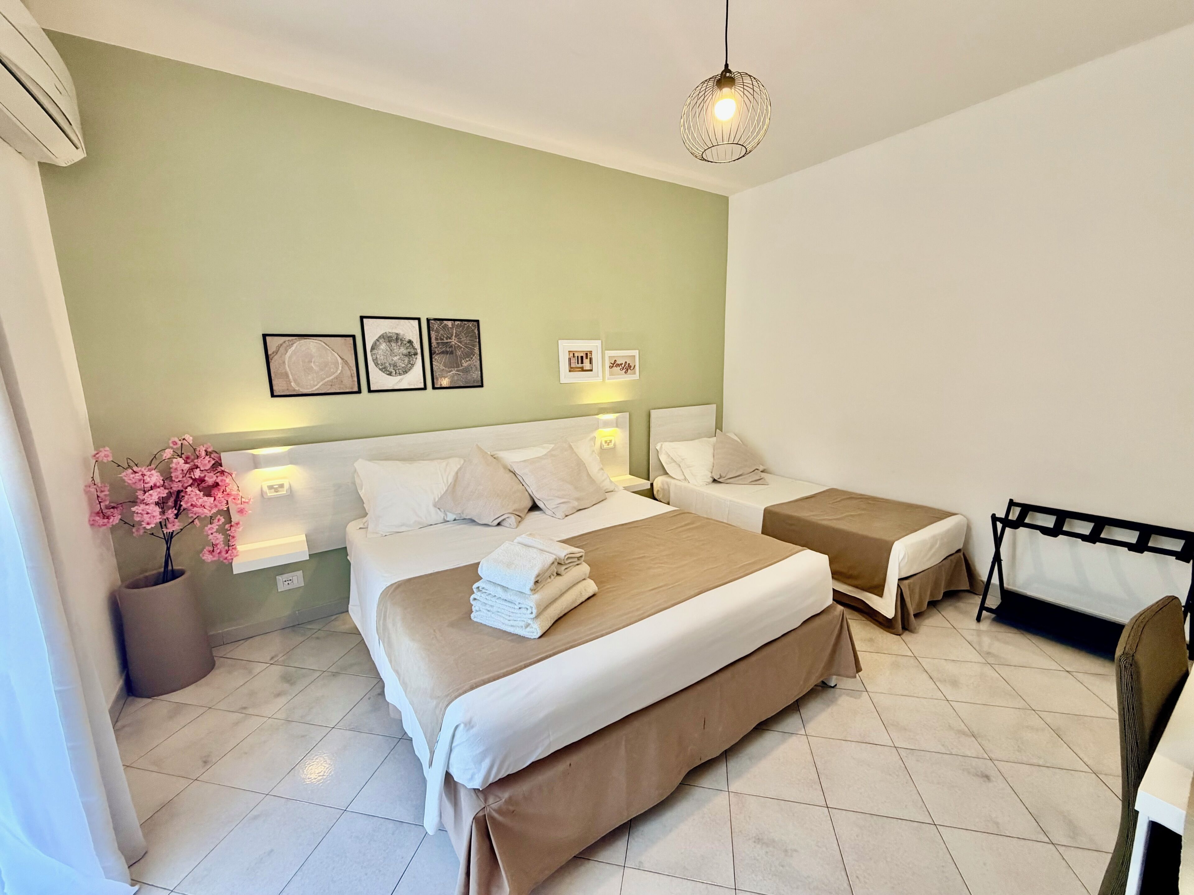 A hotel room with two beds, a potted plant, artwork on the wall, and a hanging light fixture.