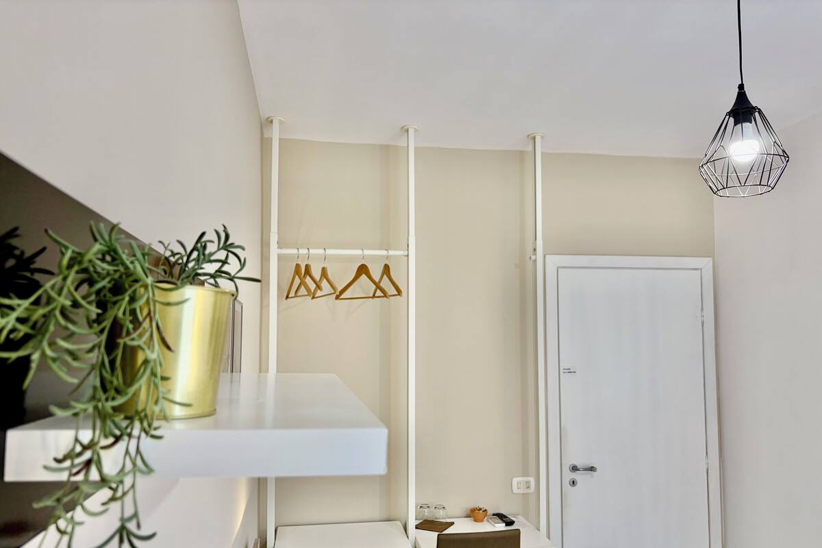 Una habitación con una puerta blanca, una lámpara colgante, un estante blanco con una planta y una mesa blanca con una silla.