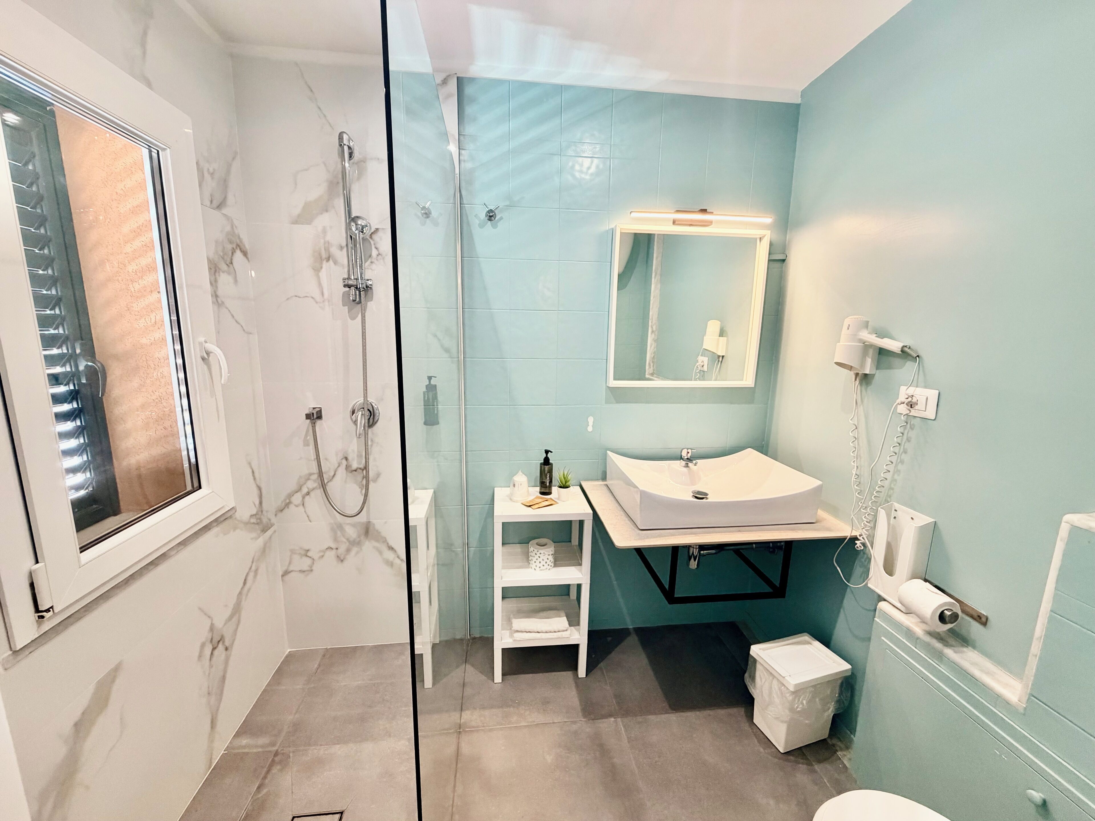 Casa de banho com cabine de chuveiro em vidro, lavatório branco, espelho e secador de cabelo na parede.