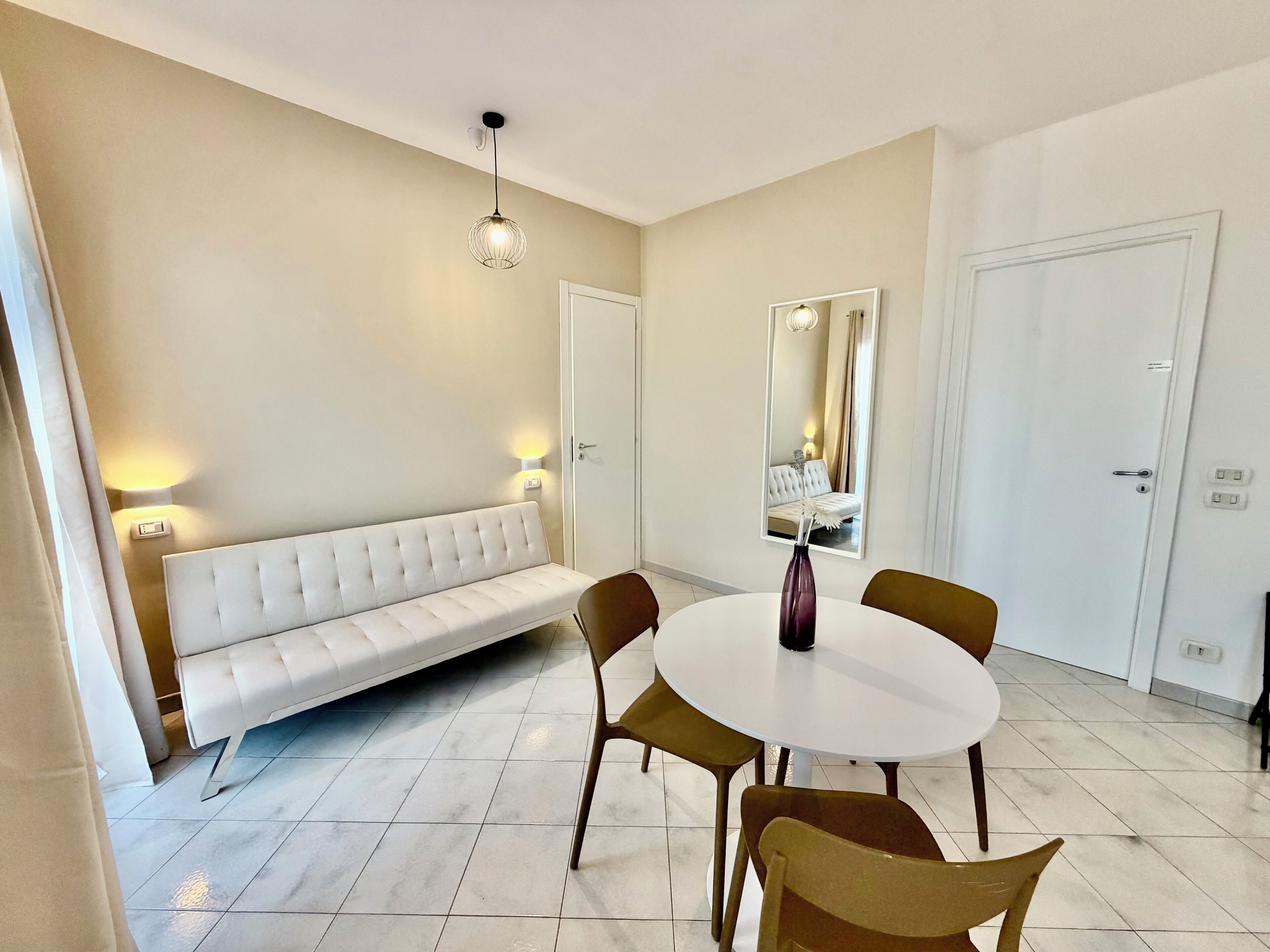 Une pièce moderne avec un canapé blanc, une table de salle à manger ronde entourée de chaises, un vase, un miroir et un luminaire au plafond.