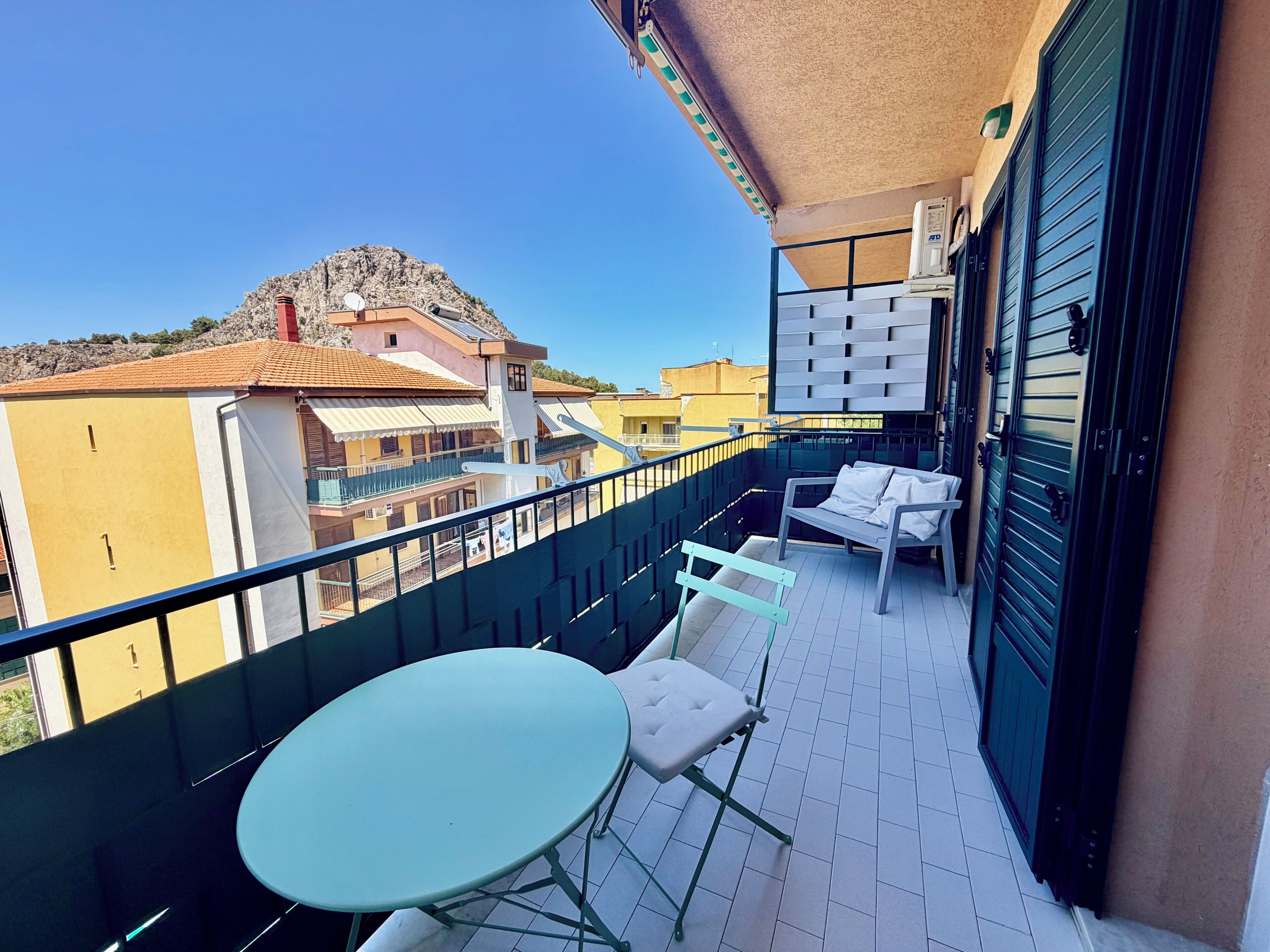A balcony with a table and chairs, a view of buildings, and a mountain in the background.