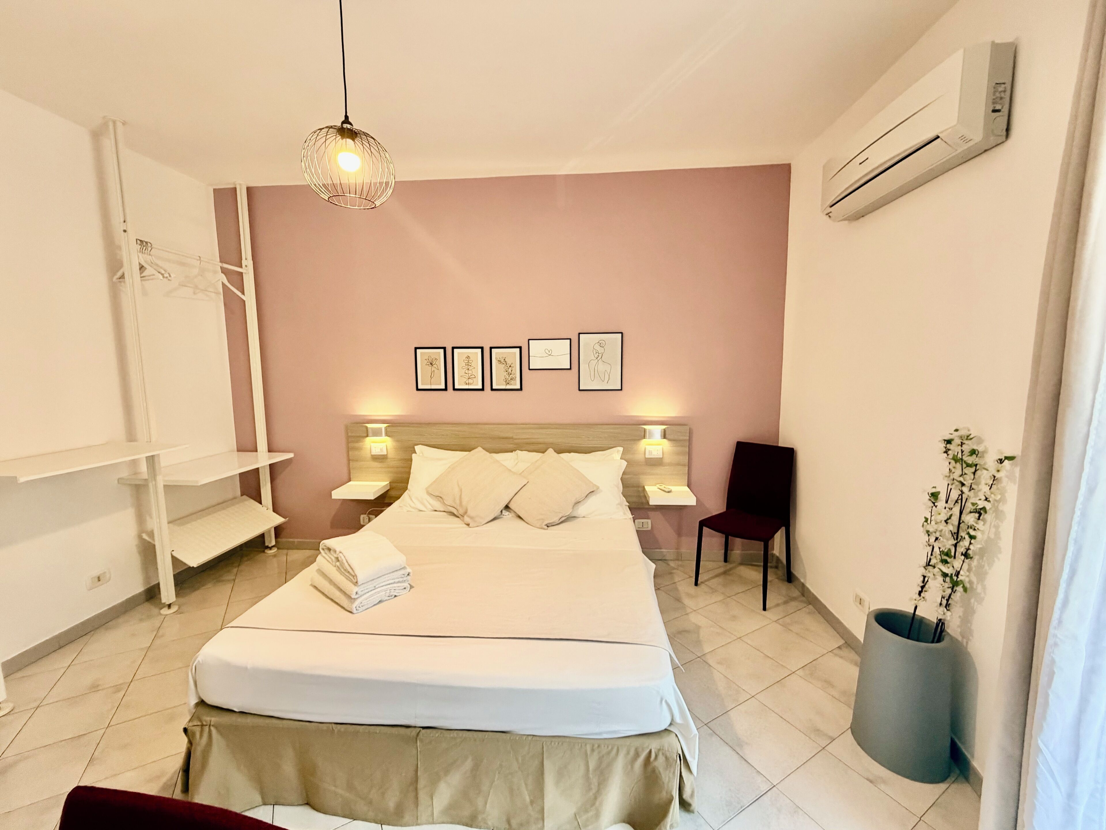 A hotel room with a bed, bedside tables, a hanging light fixture, wall-mounted air conditioning, and a potted plant.