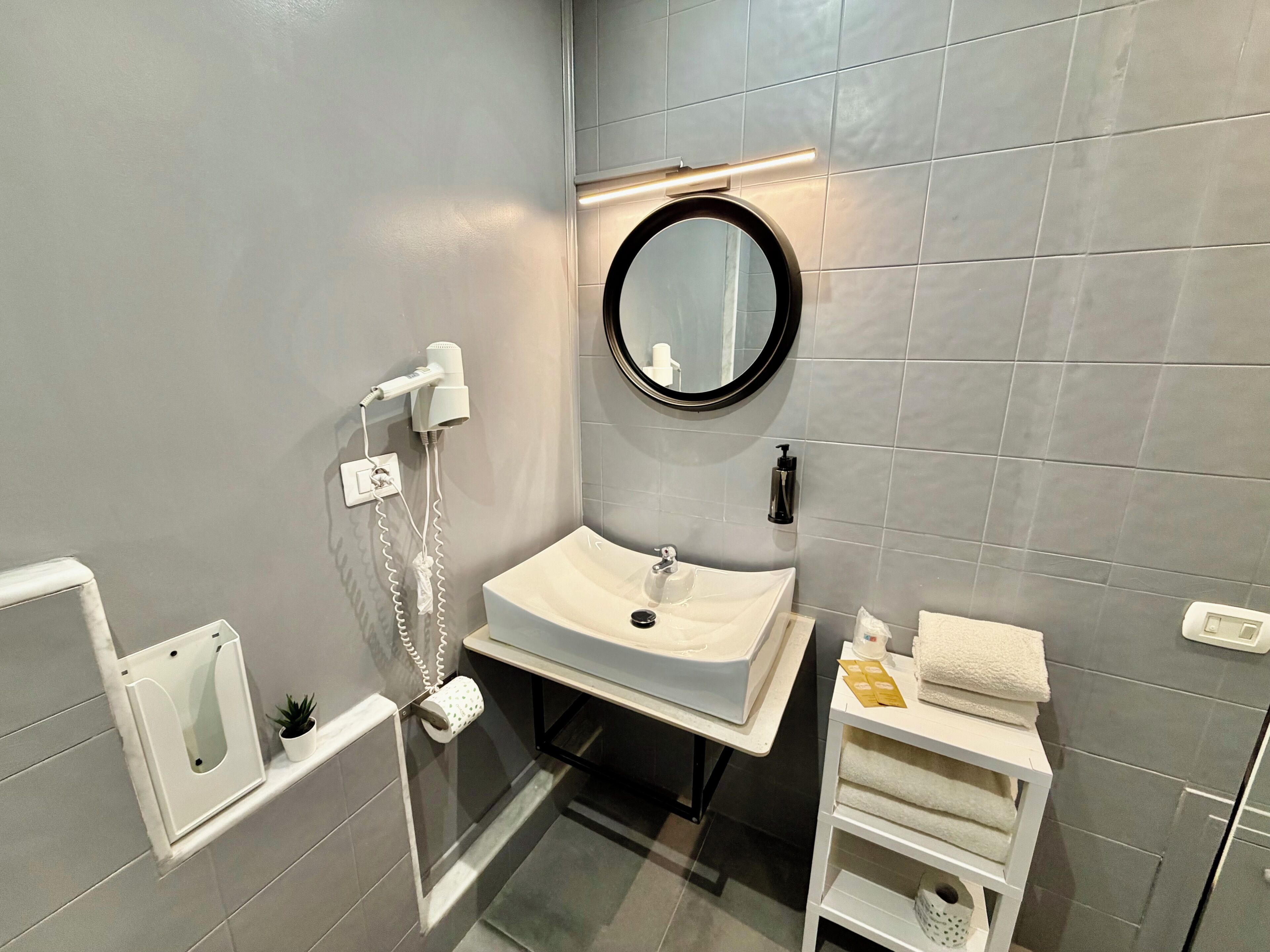 Un baño moderno con un lavabo blanco, un espejo redondo, un perchero para toallas y un secador de pelo.