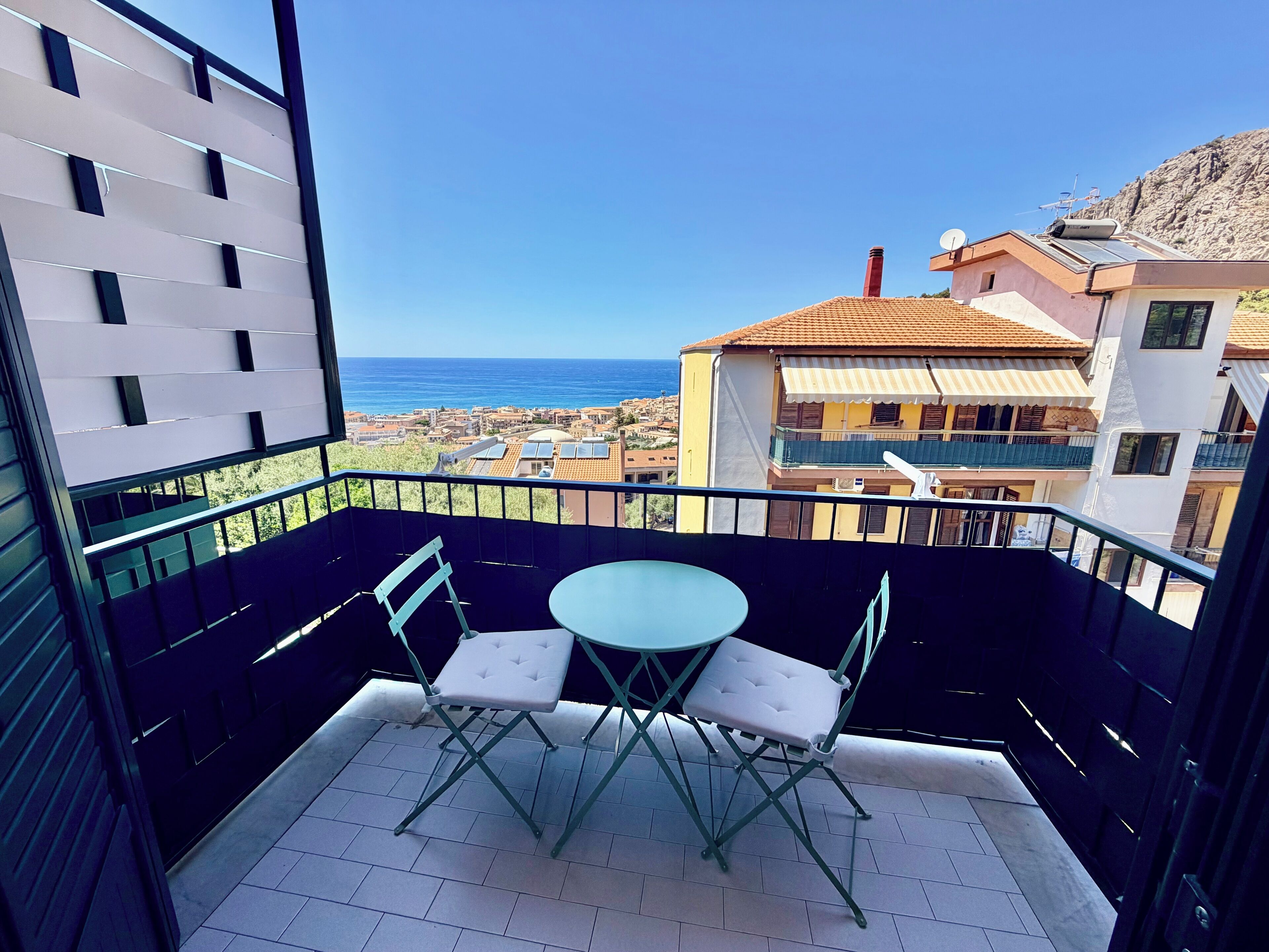A balcony with a table and chairs, overlooking a coastal town and the sea.
