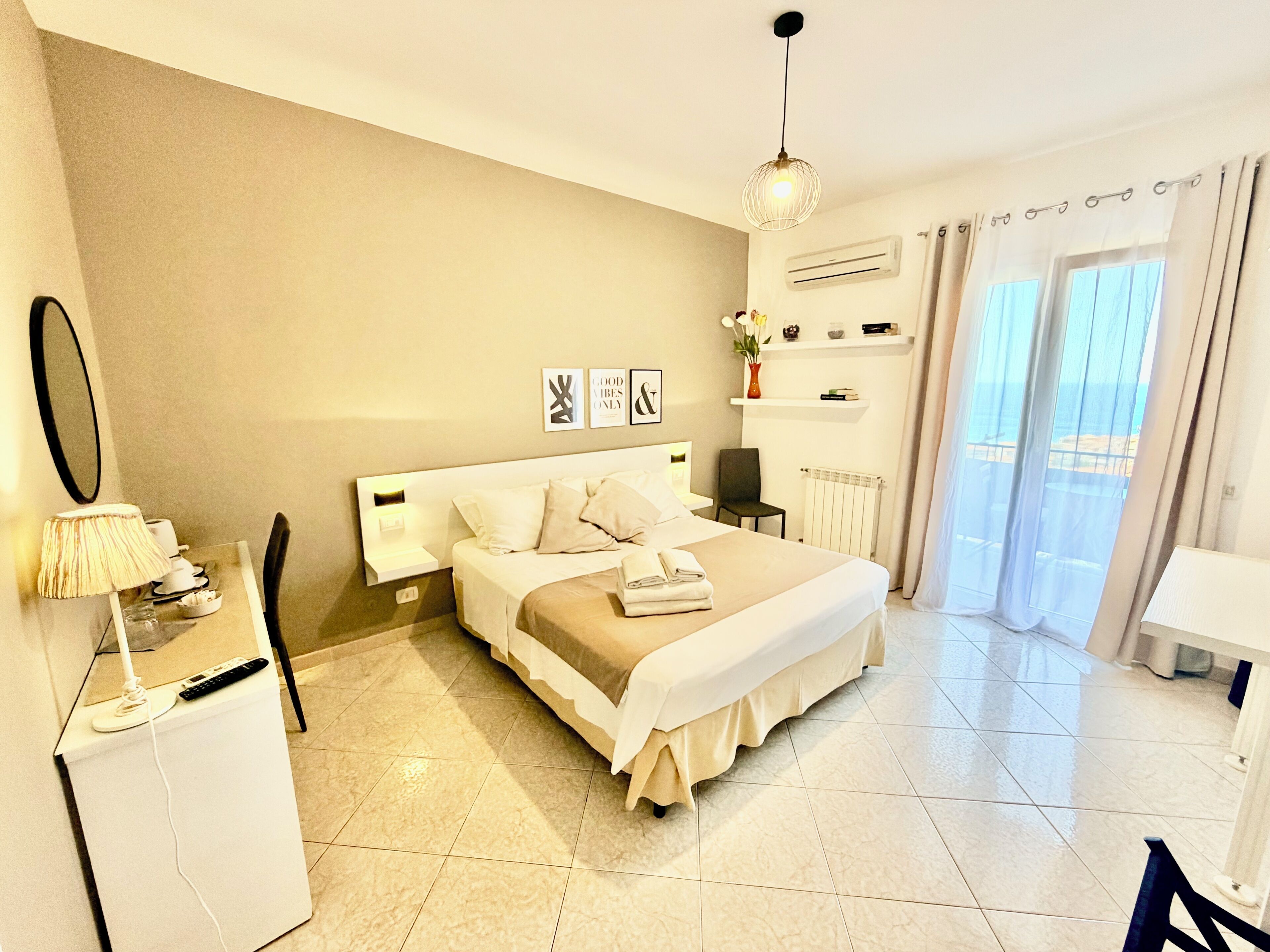Habitación de hotel con cama, mesitas de noche, escritorio, silla, lámpara, espejo y vista al mar por la ventana.