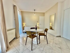 Ein Essbereich mit rundem Tisch, vier Stühlen, einem weißen Sofa, einem Heizkörper, einer Wandleuchte und einem Spiegel.