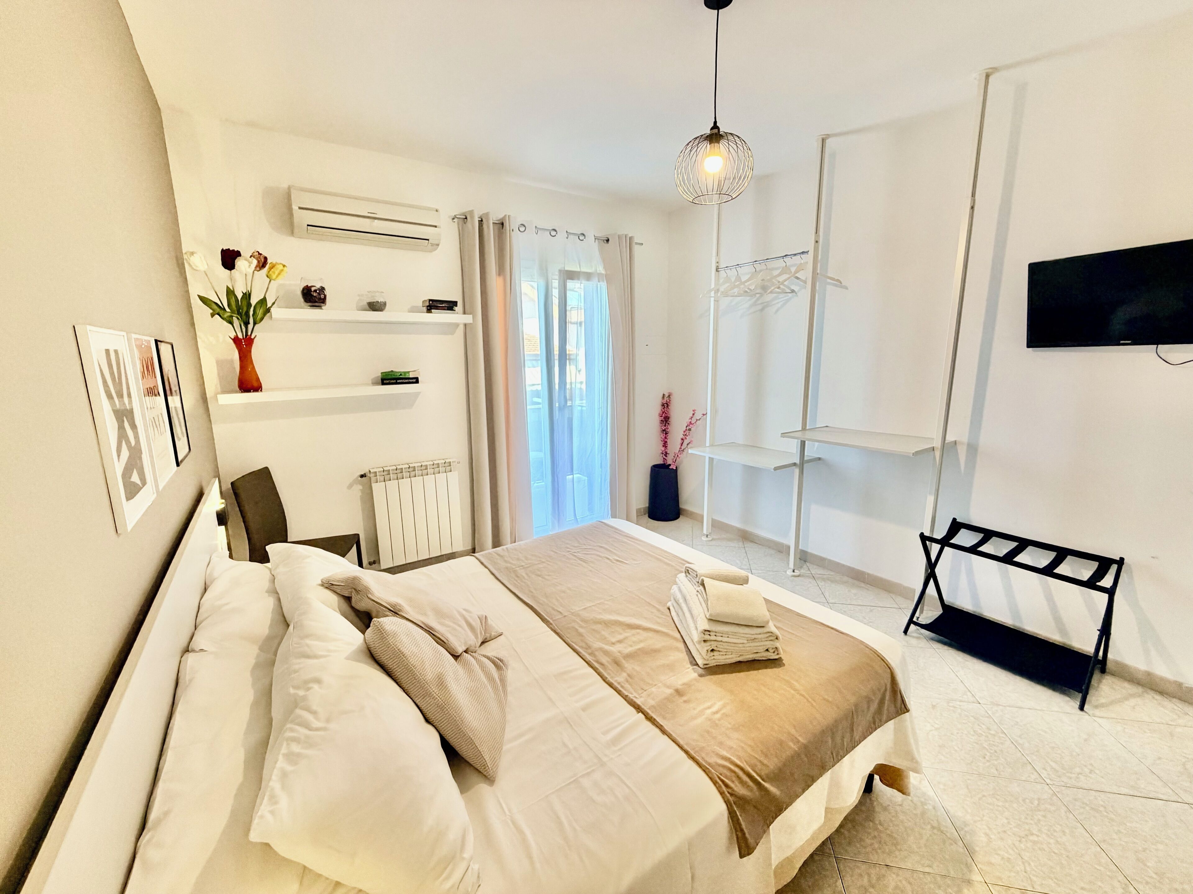 A modern bedroom with a bed, a TV, a radiator, a shelf with decorative items, and a window with curtains.
