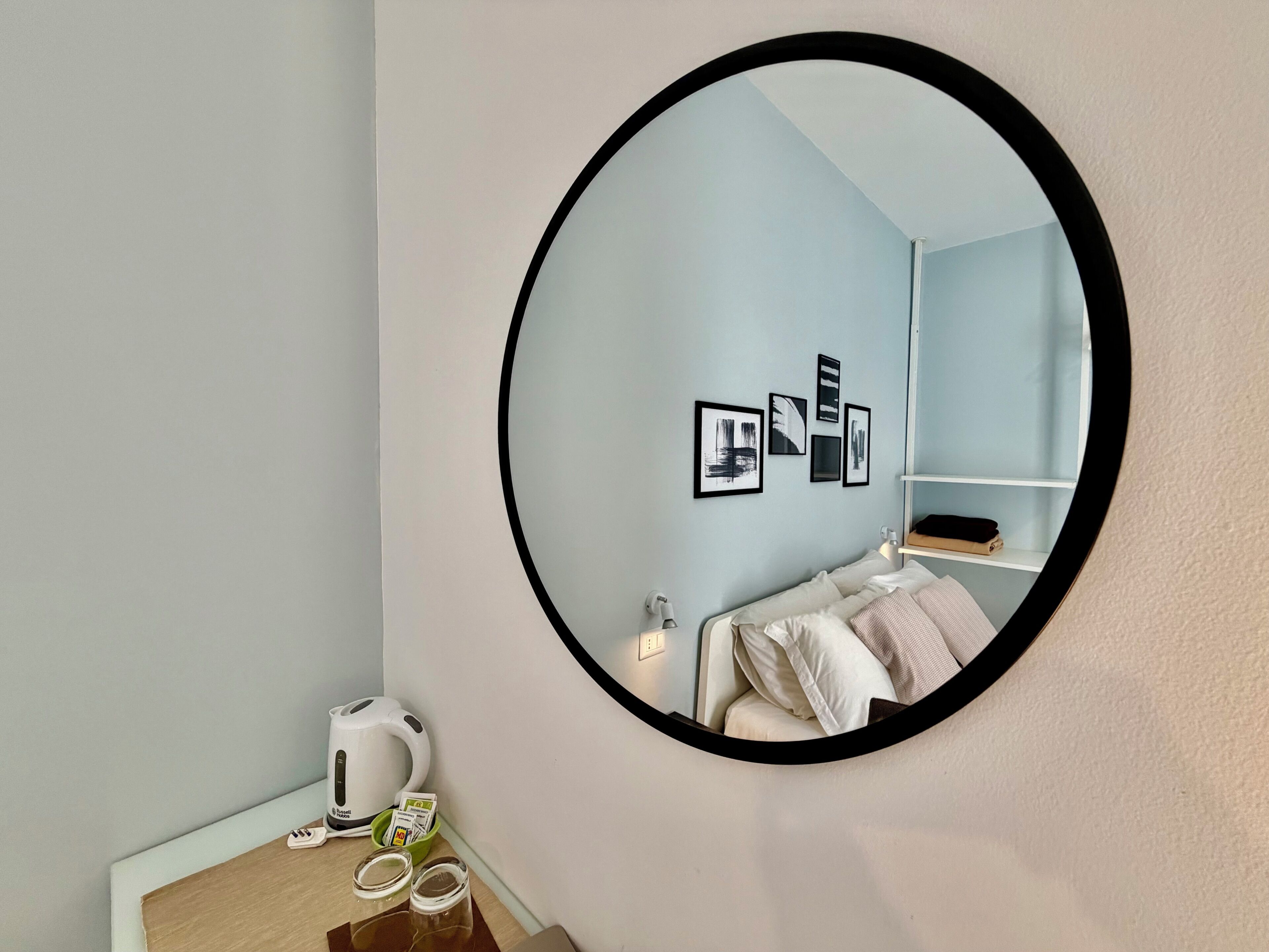 A room with a round mirror, a bed with pillows, a small table with a kettle and tea bags, and framed pictures on the wall.
