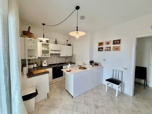 Ett modernt kök med vita skåp, en köksö, en matplats med en stol och en vägg med inramade tavlor.