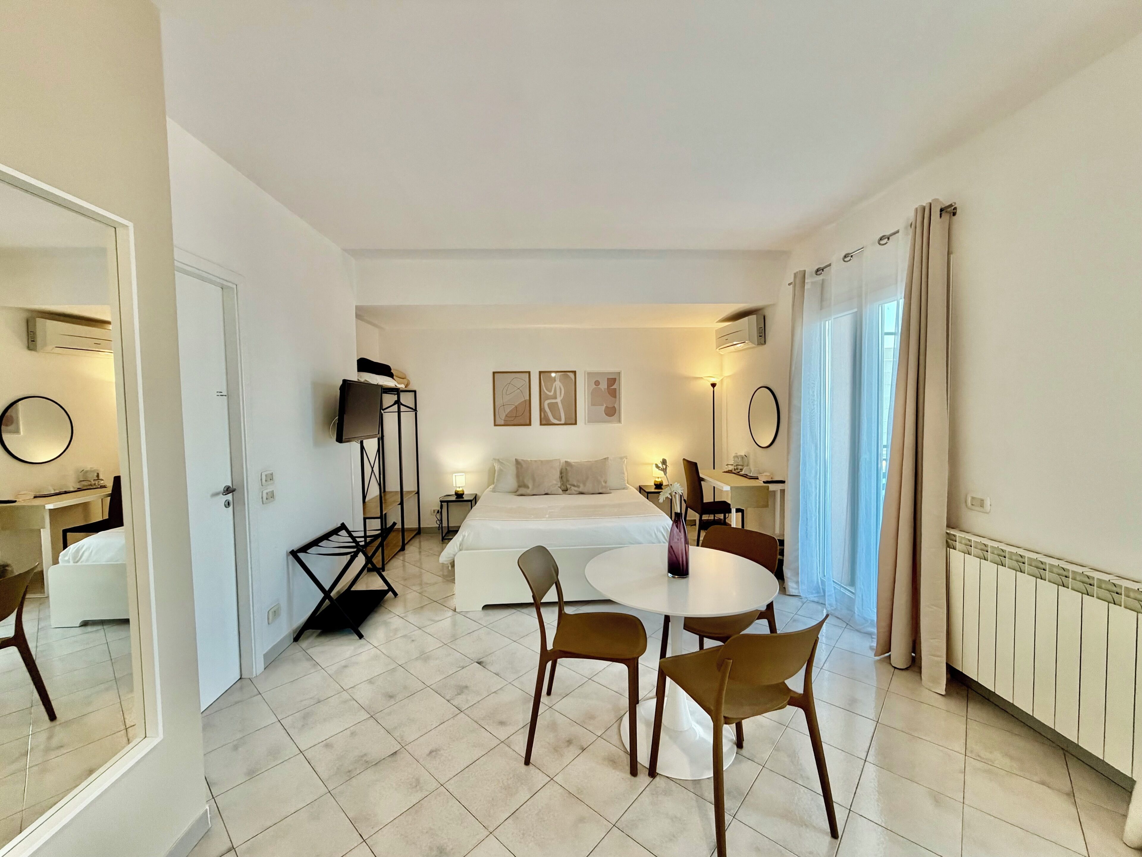 A hotel room with a bed, a desk, a TV, and a dining area with chairs.